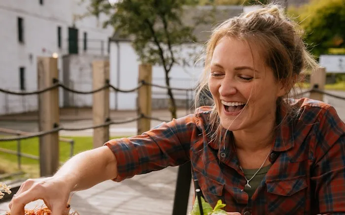 Katie Tunn, a blonde woman, sits outside at a wooden table laden with plates and bowls of food. She reaches for food on a plate while smiling.