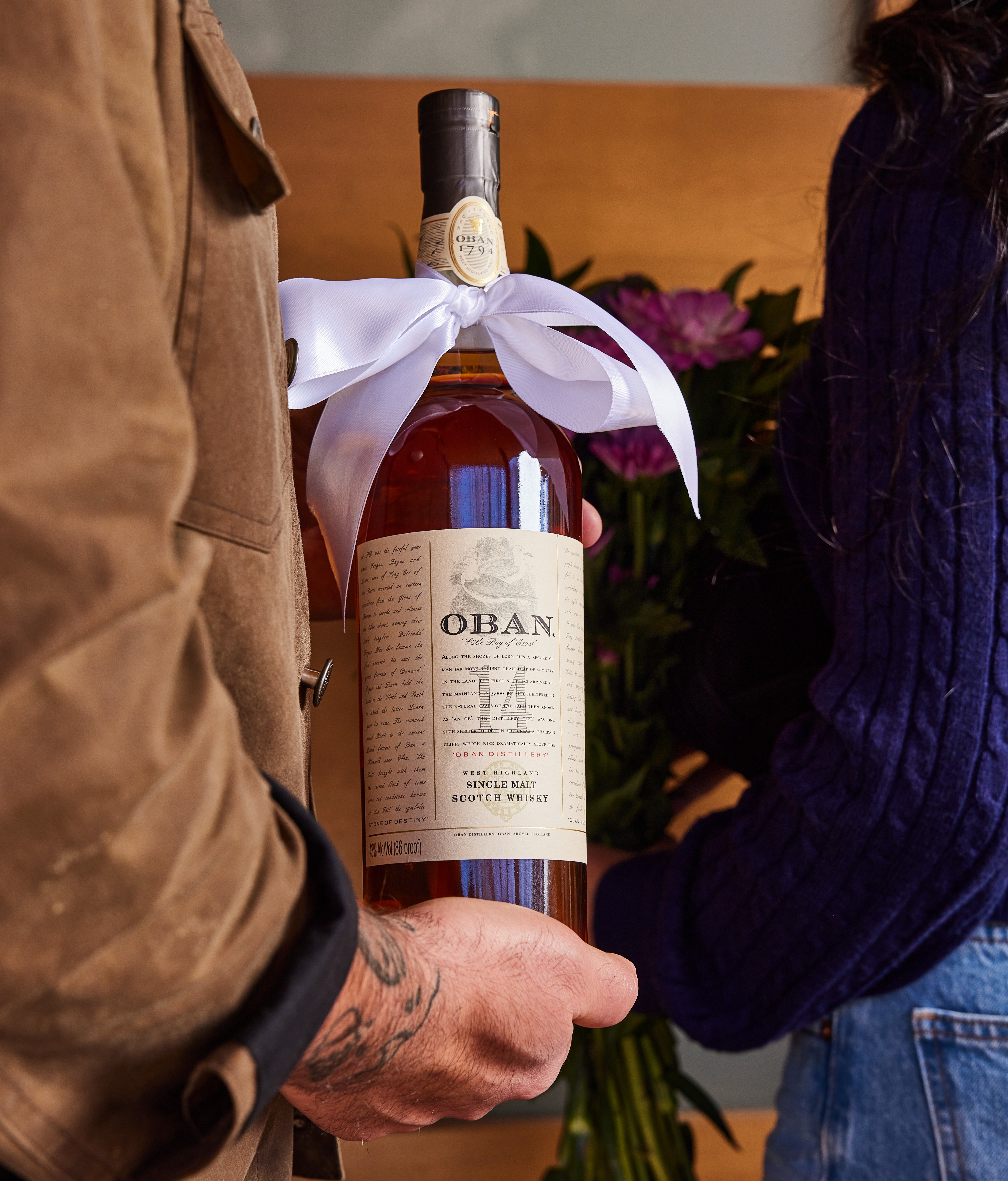 A man holds a bottle of Oban 14 year old whisky with a white bow tied around the neck. A woman stands next to him holding a bouquet of flowers.