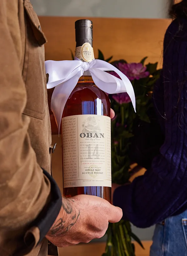 Ein Mann hält eine Flasche Oban 14 Jahre alten Whisky mit einer weißen Schleife um den Hals gebunden. Neben ihm steht eine Frau mit einem Blumenstrauß.