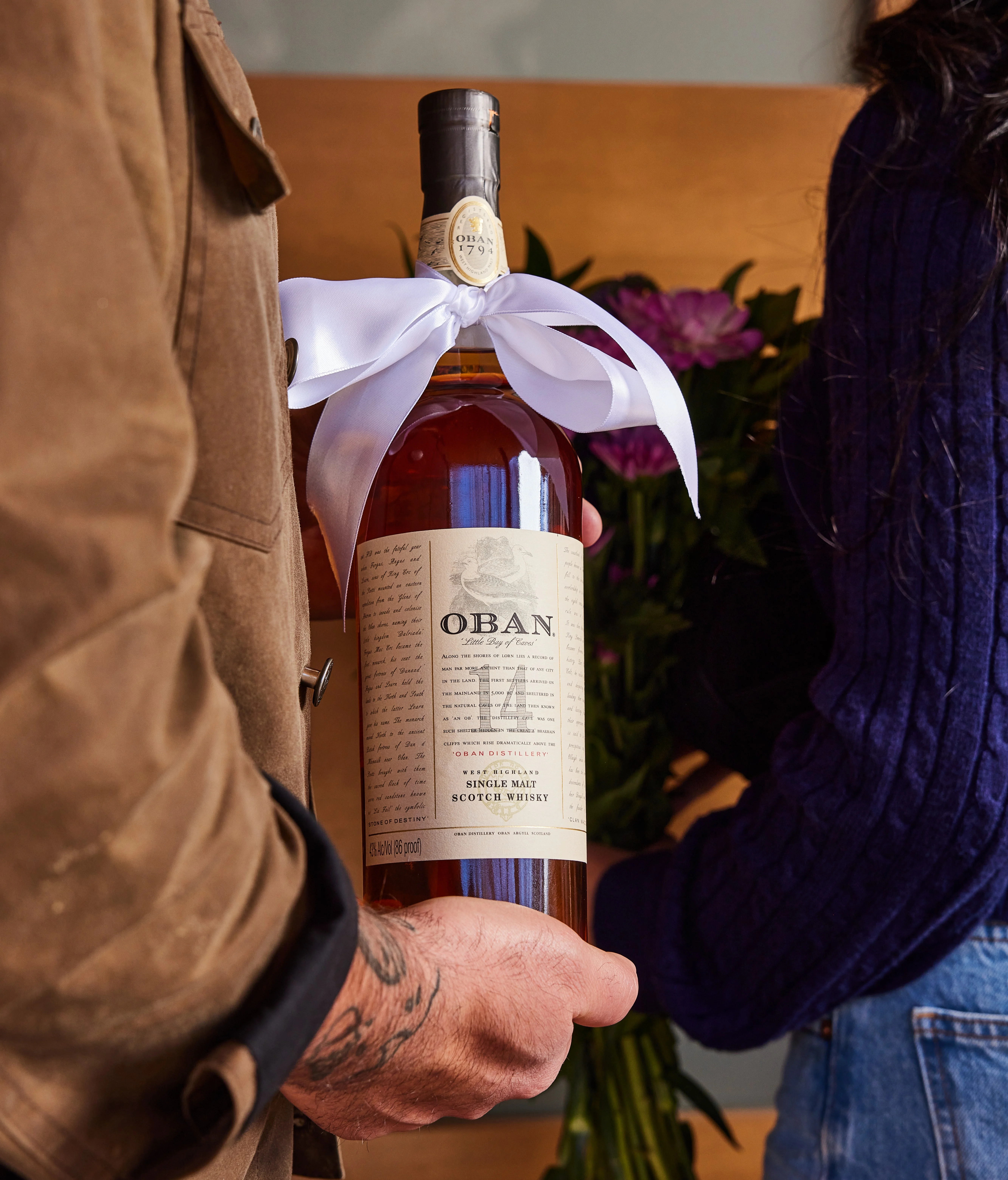 A man holds a bottle of Oban 14 year old whisky with a white bow tied around the neck. A woman stands next to him holding a bouquet of flowers.