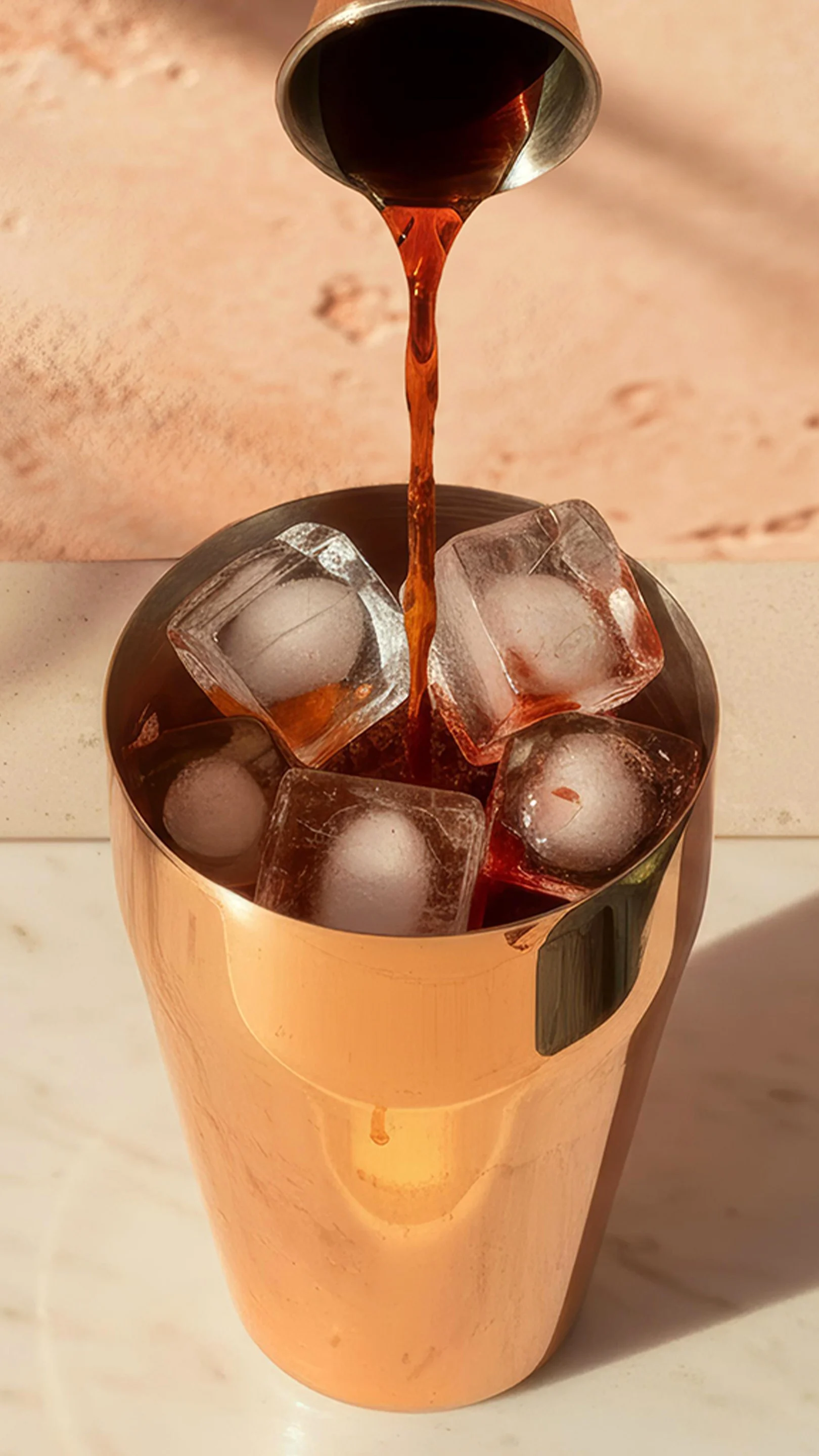 A brown liquid is poured into a copper cocktail shaker over ice, with a bowl of green olives nearby