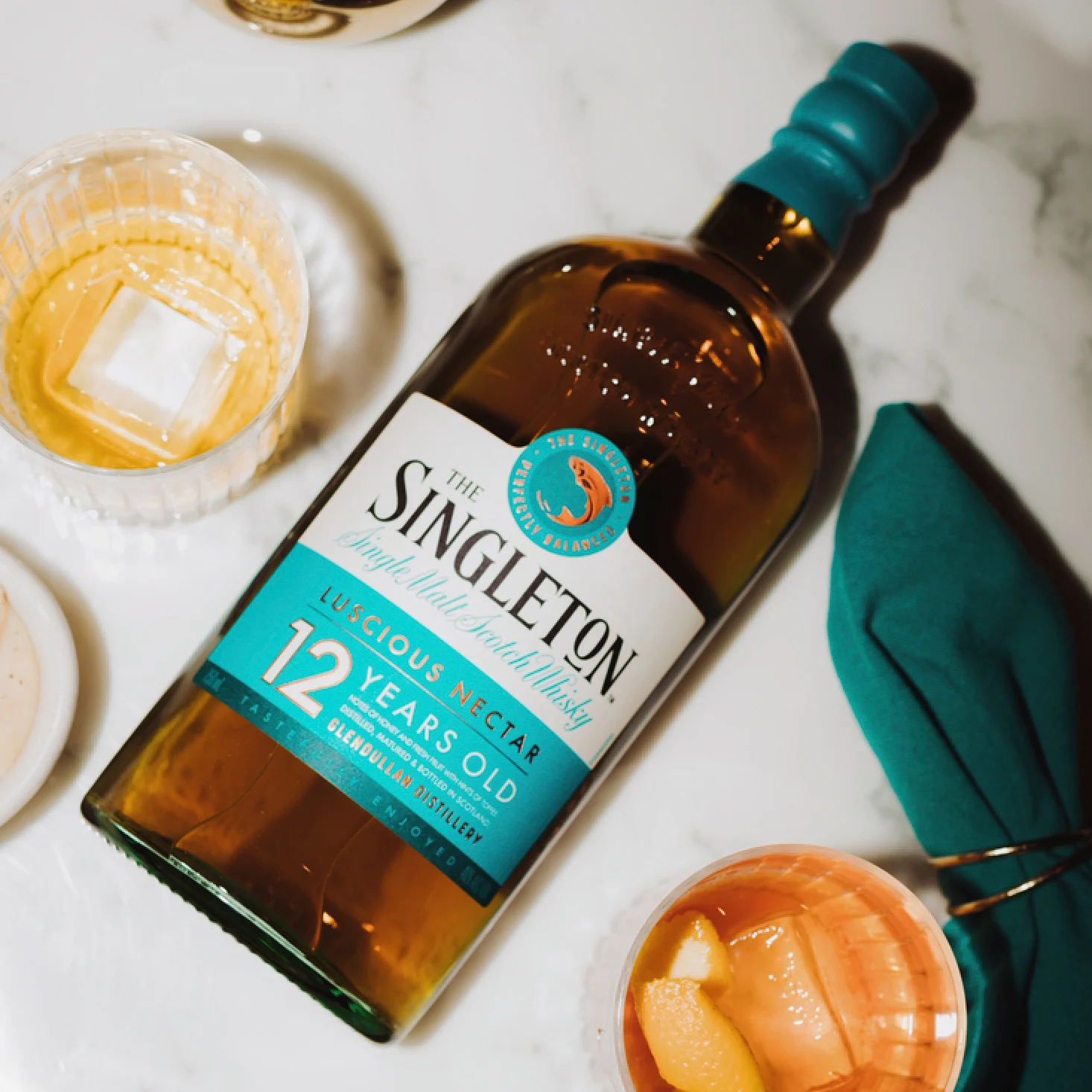 The Singleton of Dufftown 12 Year Old bottle lying on a marble surface with two whiskey glasses and a teal napkin