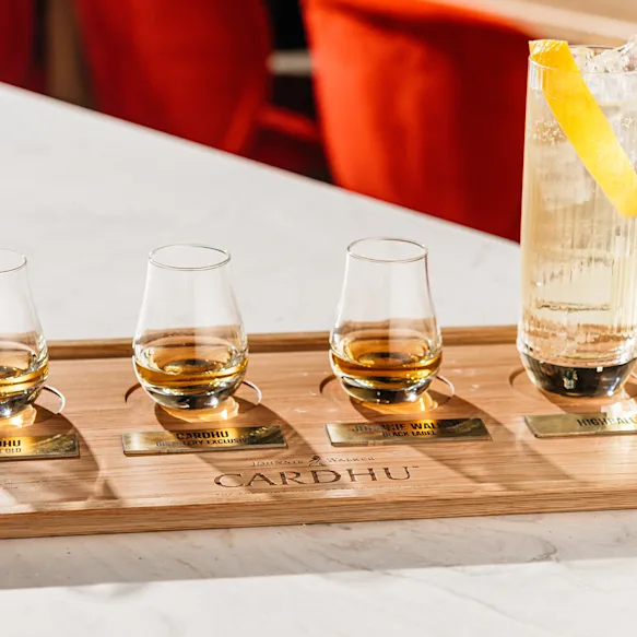 Three small whisky glasses one third full and a highball cocktail displayed on top of a wooden board written Cardhu