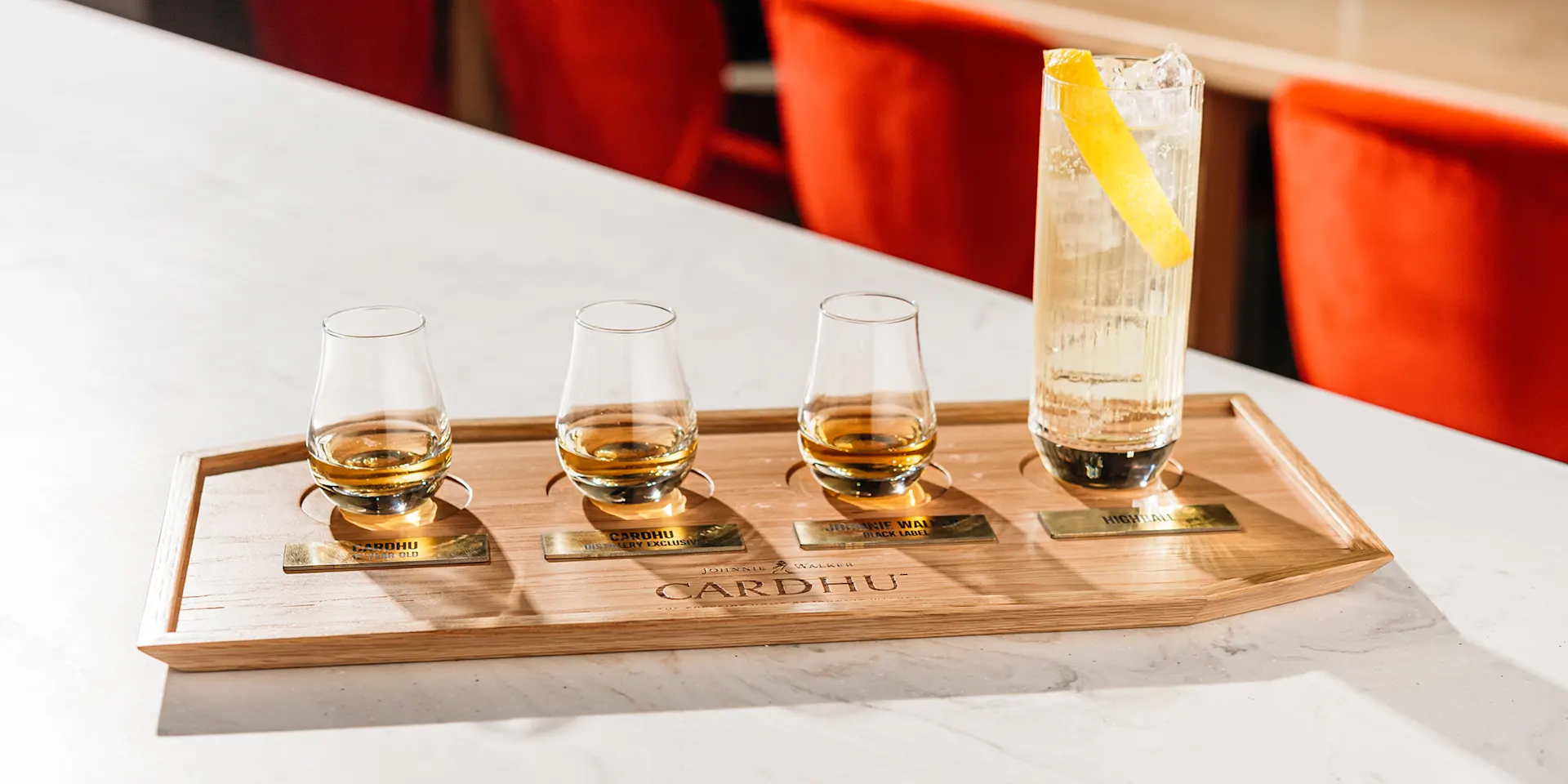 Three small whisky glasses one third full and a highball cocktail displayed on top of a wooden board written Cardhu