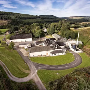 A white building with a grey roof is pictured from up high. Surrounding the building are hills featuring grasses and heather. Behind the building are higher hills with a forest of green trees.