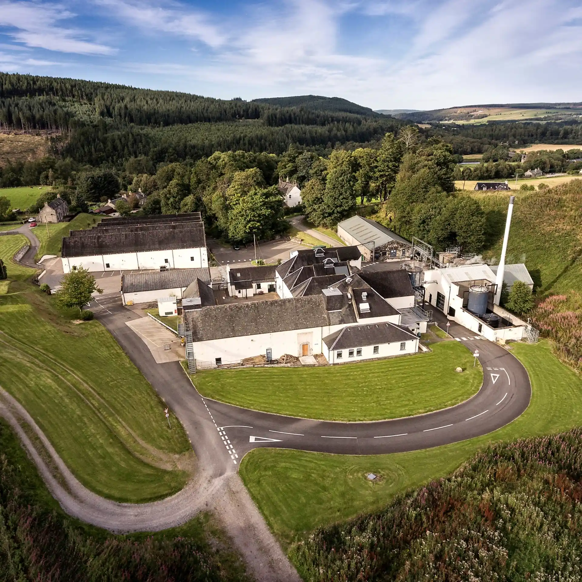 A white building with a grey roof is pictured from up high. Surrounding the building are hills featuring grasses and heather. Behind the building are higher hills with a forest of green trees.