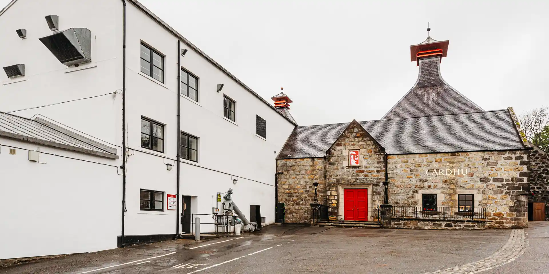 The brown stone building of the Cardhu distillery sits next to a white building. The building has a pitched slate roof and a red door. Attached to the brown stone wall is a gold metal sign with the words ‘Cardhu’.