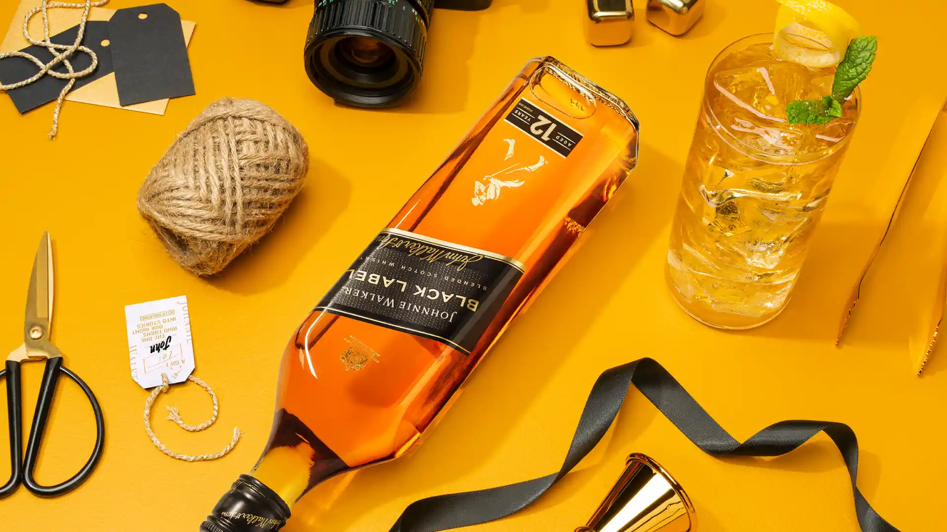 Eine Flasche Johnnie Walker Black Label liegt auf einem gelben Tisch, daneben liegen Schnur, Schere und ein langer Whisky-Cocktail.