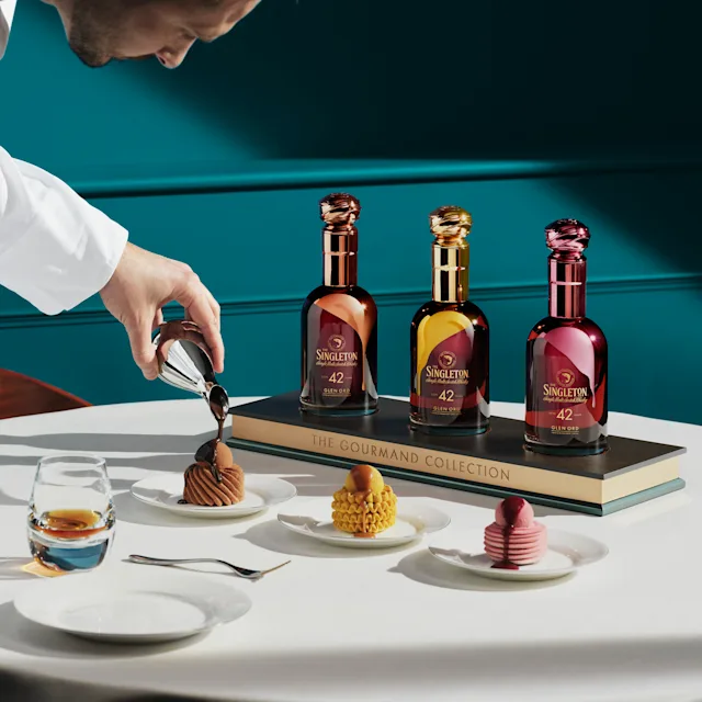Chef Nicolas Rouzaud pouring sauce over a dessert, with The Singleton Gourmand Collection bottles displayed alongside three desserts and a glass of whiskey.
