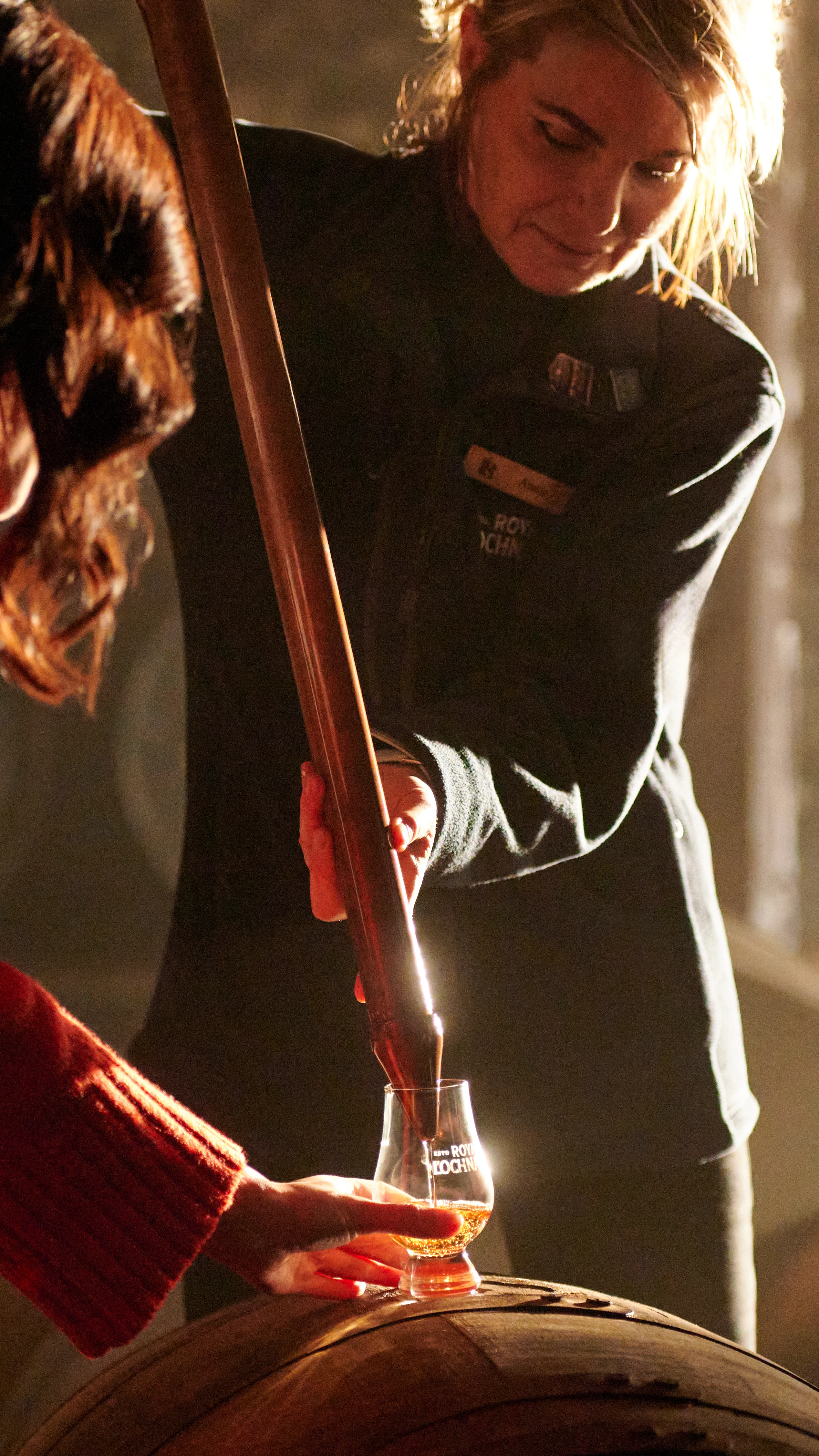 A woman draws whisky from a wooden barrel using a metal pipette. She pours the whisky into a tasting glass held by a woman in a red jumper, who holds the glass on top of the barrel.