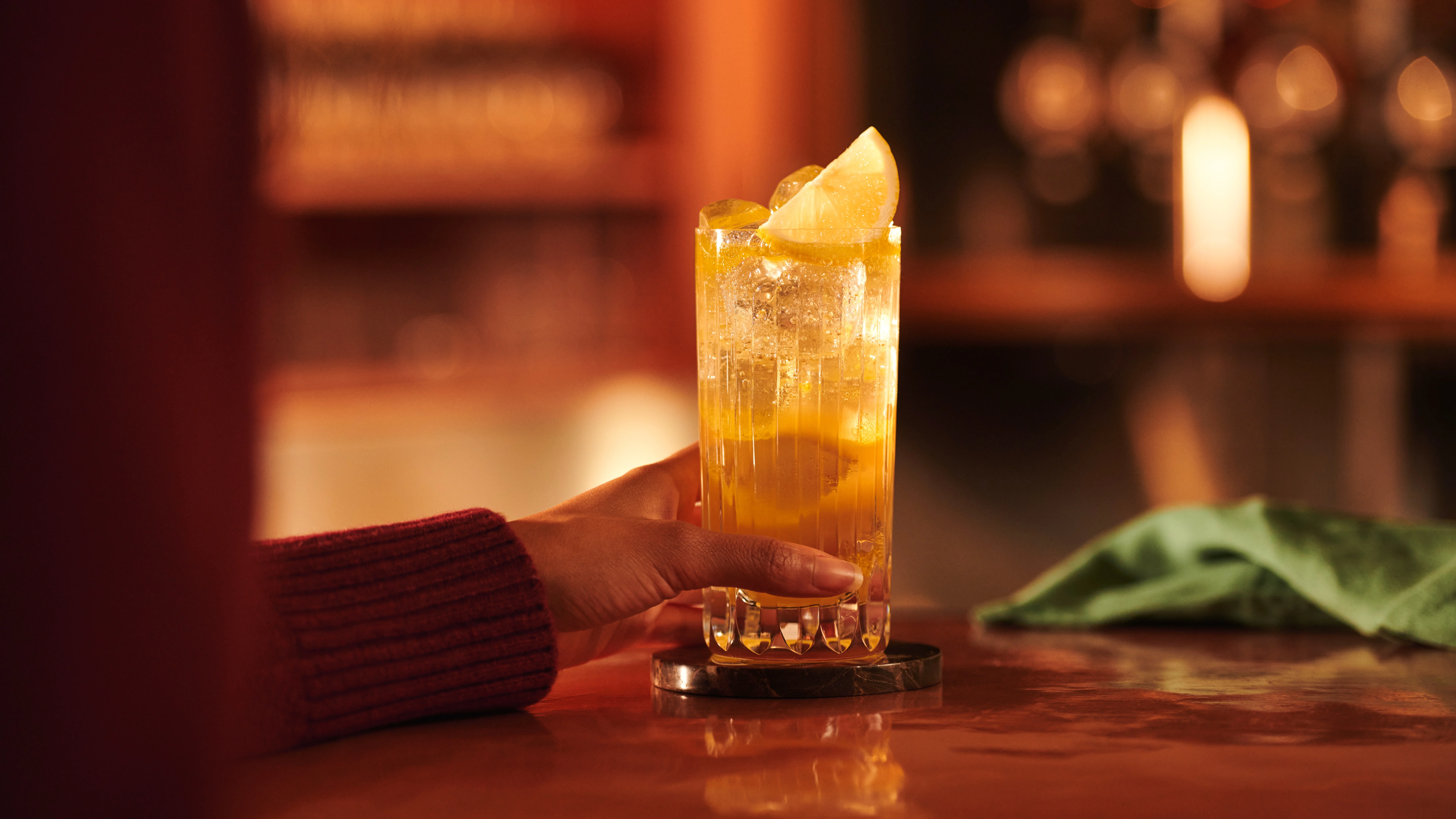 A woman places a hand round a whisky highball cocktail in a highball glass while sat at a copper bar