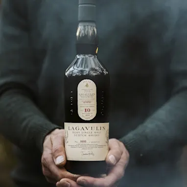 Twee kleine boten liggen op het water voor een wit gebouw met de woorden Lagavulin op de muur geschilderd. Naast het gebouw liggen met gras begroeide heuvels; de lucht is blauw en bezaaid met witte wolken.