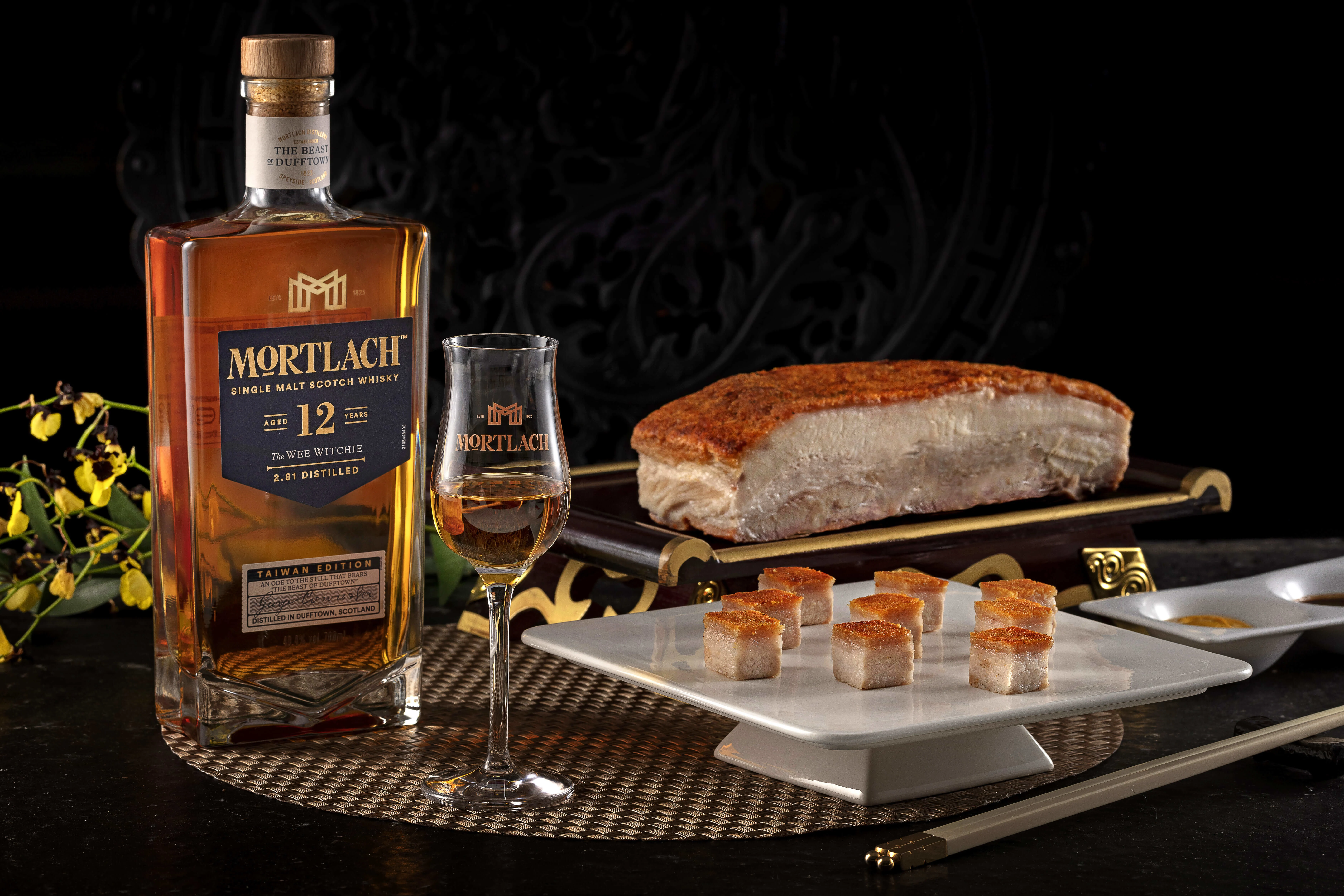 一瓶和一杯 Mortlach 12 年的 The Wee Witchie 放在桌上,旁邊有切片和方塊狀的豬五花。