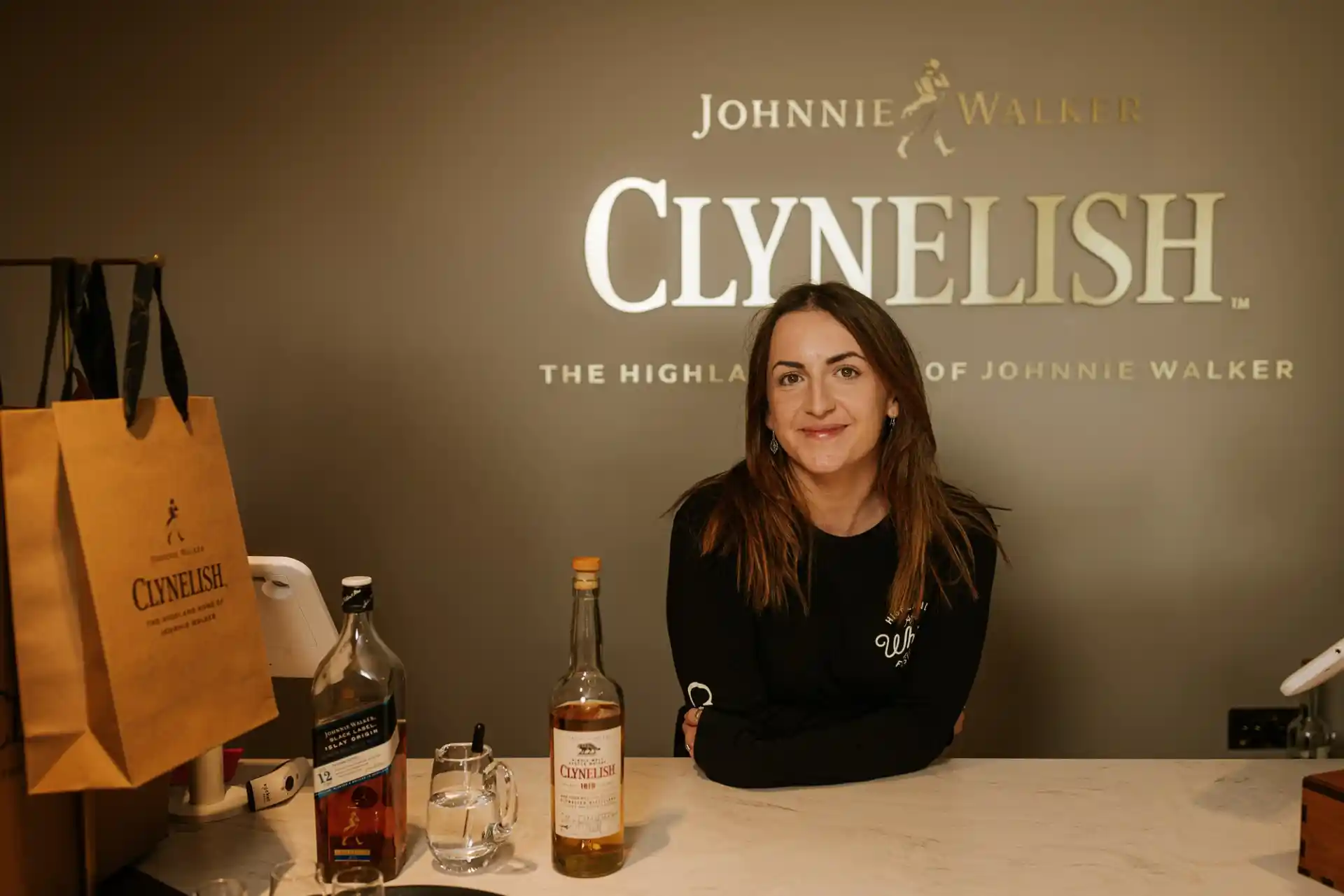 Een glimlachende Clynelish-medewerker leunt achter een toonbank waarop een Clynelish-cadeauzakje, twee whiskyflessen en een waterkan staan. Achter de medewerker, op de muur, staan de woorden 'Clynelish, the highland home of Johnnie Walker'.