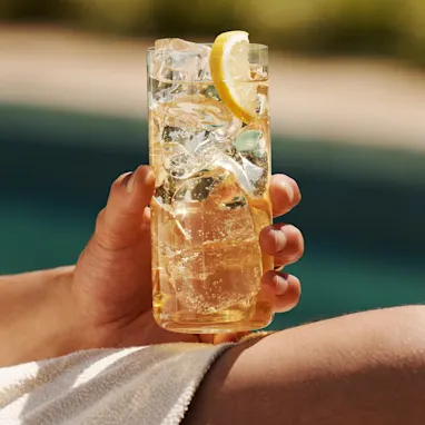 A man holds a whisky highball on his knee while sat next to a pool