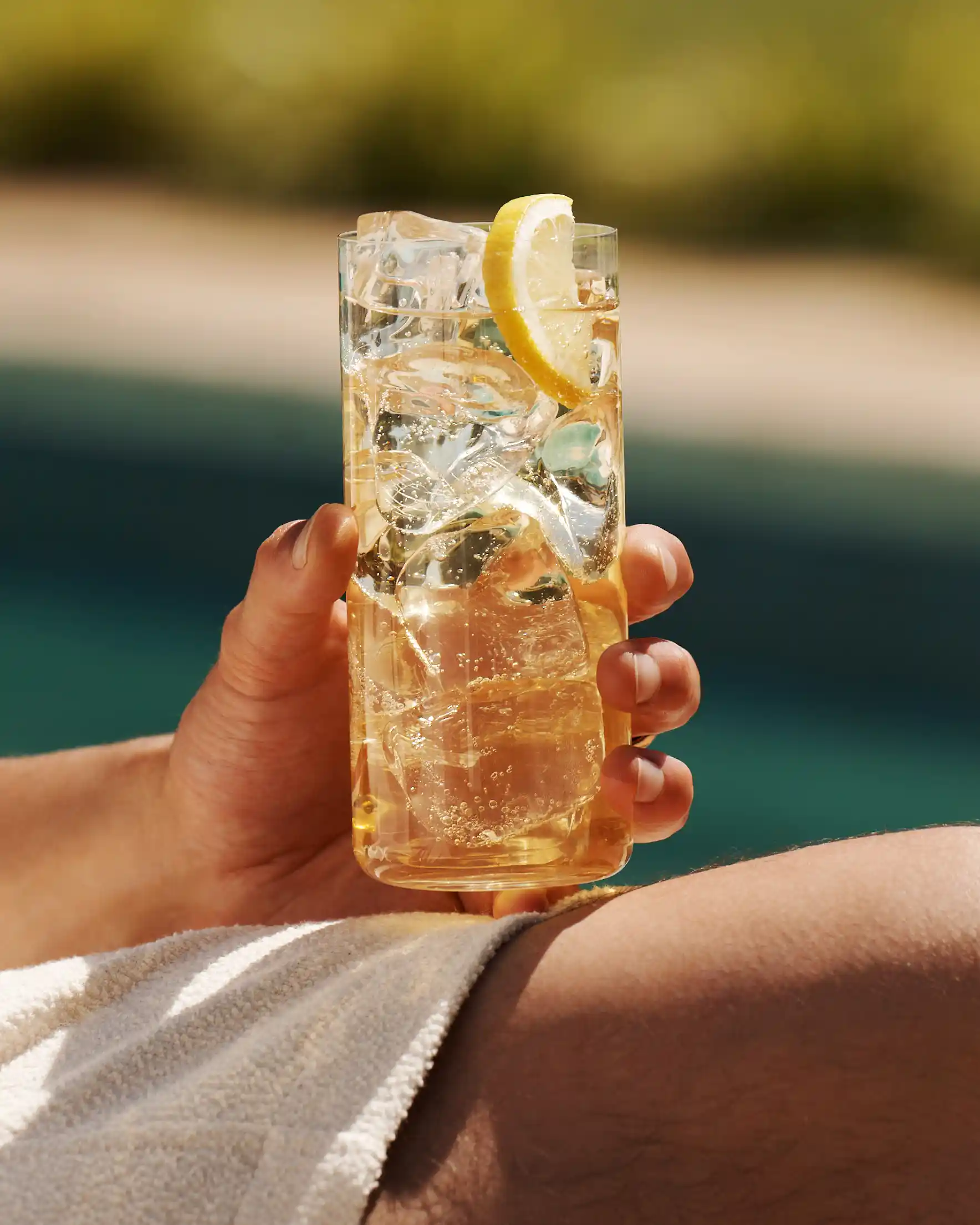 A man holds a whisky highball on his knee while sat next to a pool