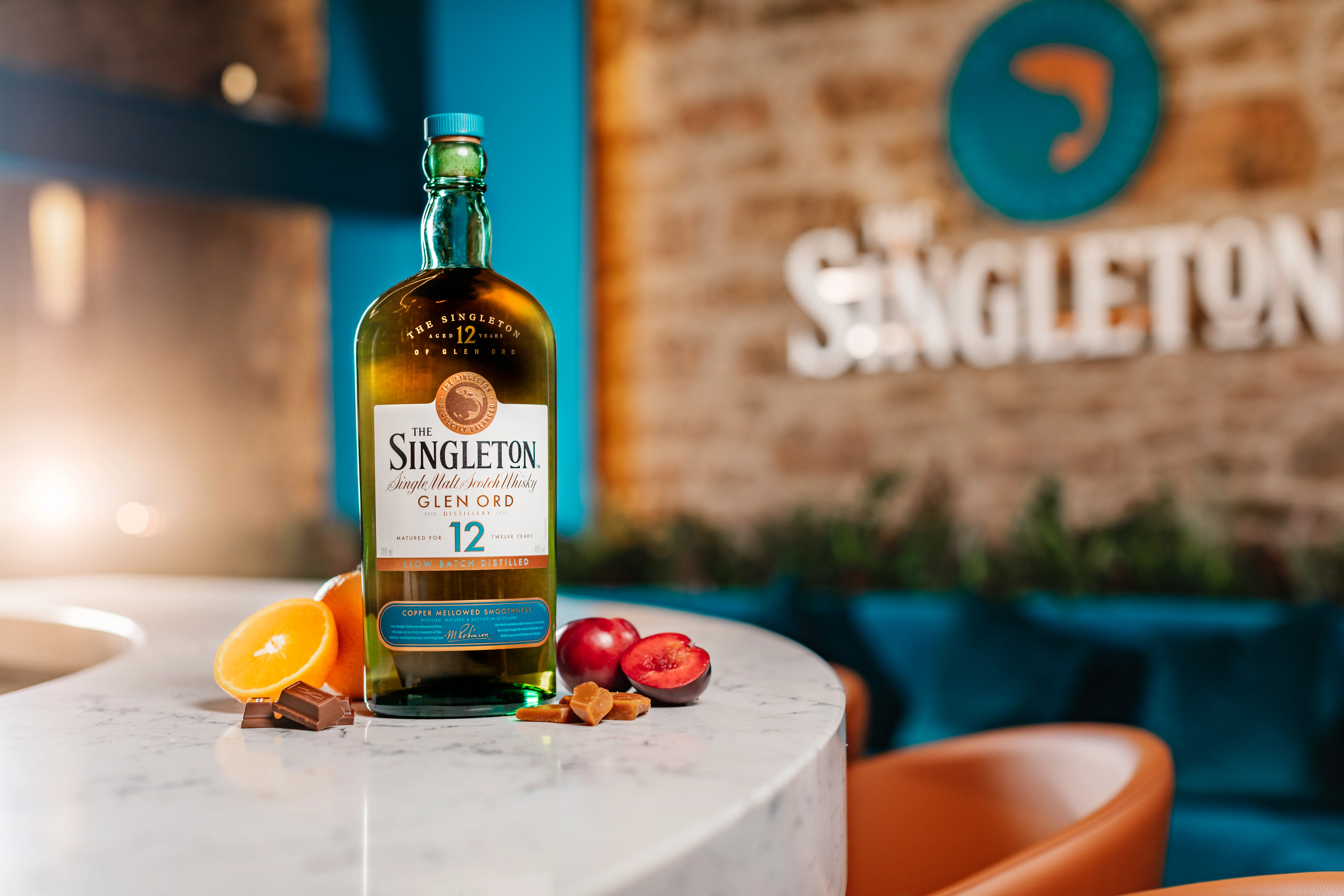 The Singleton Glen Ord 12 year old bottle is sitting on a counter in The Singleton distillery surrounded by fruits used to create the liquid.
