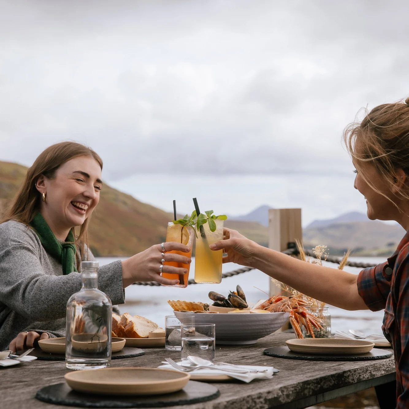 Two friends clink glasses as they eat dinner outside by a lake.