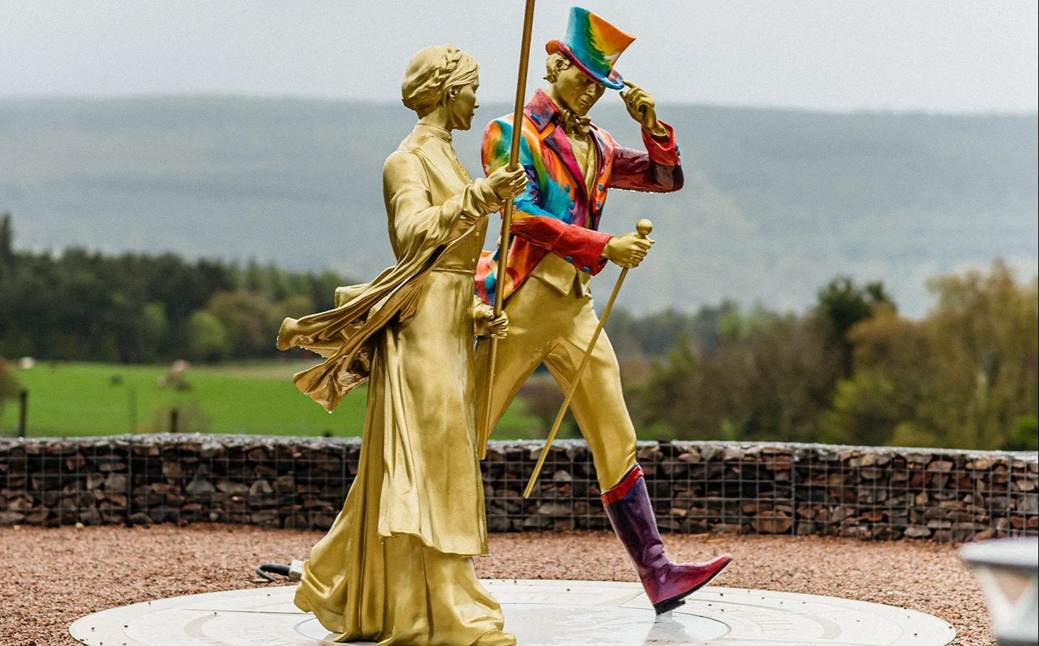 A golden statue of Helen Cummings holding a red flag stands next to a golden statue of the Johnnie Walker 'Striding Man' wearing a rainbow jacket and top hat and holding a walking cane. Both the statues appear to be walking.