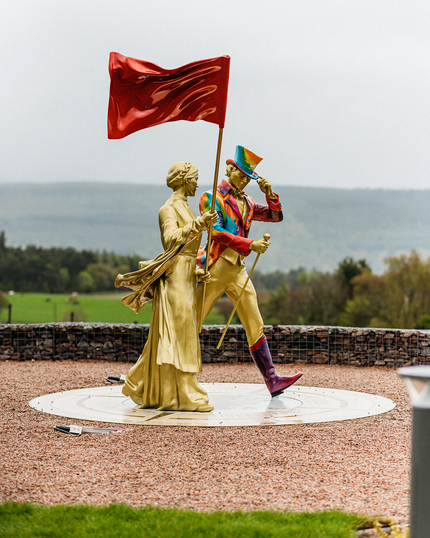 A golden statue of Helen Cummings holding a red flag stands next to a golden statue of the Johnnie Walker 'Striding Man' wearing a rainbow jacket and top hat and holding a walking cane. Both the statues appear to be walking.
