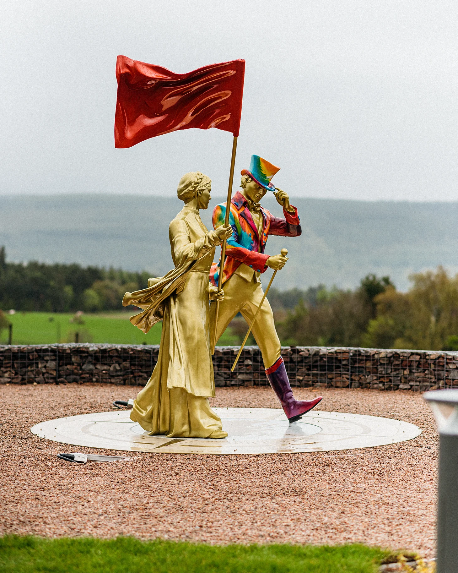 A golden statue of Helen Cummings holding a red flag stands next to a golden statue of the Johnnie Walker 'Striding Man' wearing a rainbow jacket and top hat and holding a walking cane. Both the statues appear to be walking.