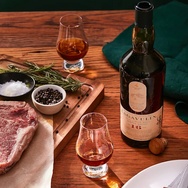 Shop Smoky Whiskies A bottle of Lagavulin 16 year old whisky sits on a wooden surface next to two dram glasses and a wooden chopping board on which there is a raw steak, a bowl of salt and pepper and a sprig of rosemary.