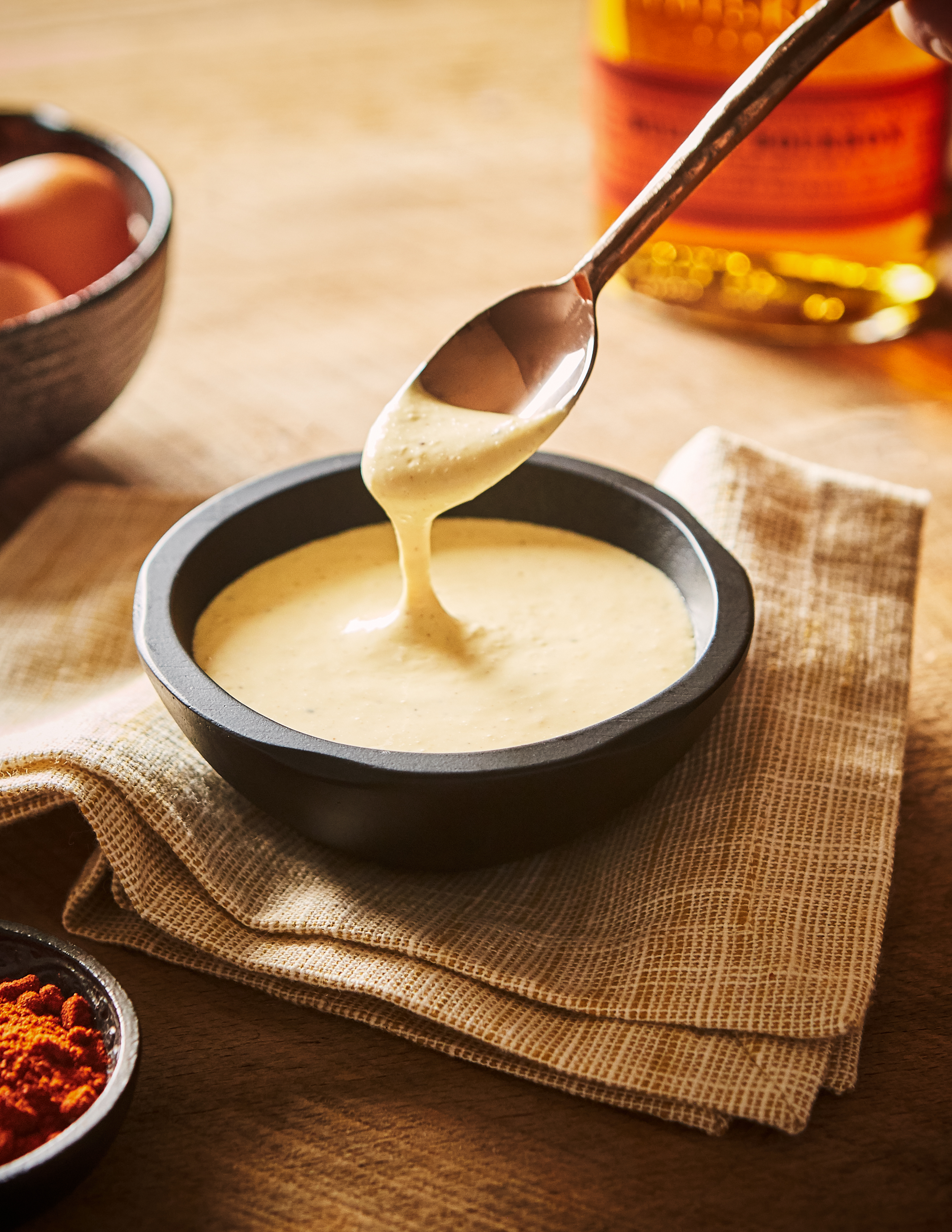 Op een tafel staat een zwarte schaal met een crèmekleurige saus. Een lepel dipt erin. Op tafel staan ook een fles bourbon, een schaaltje eieren en een schaaltje kruiden.