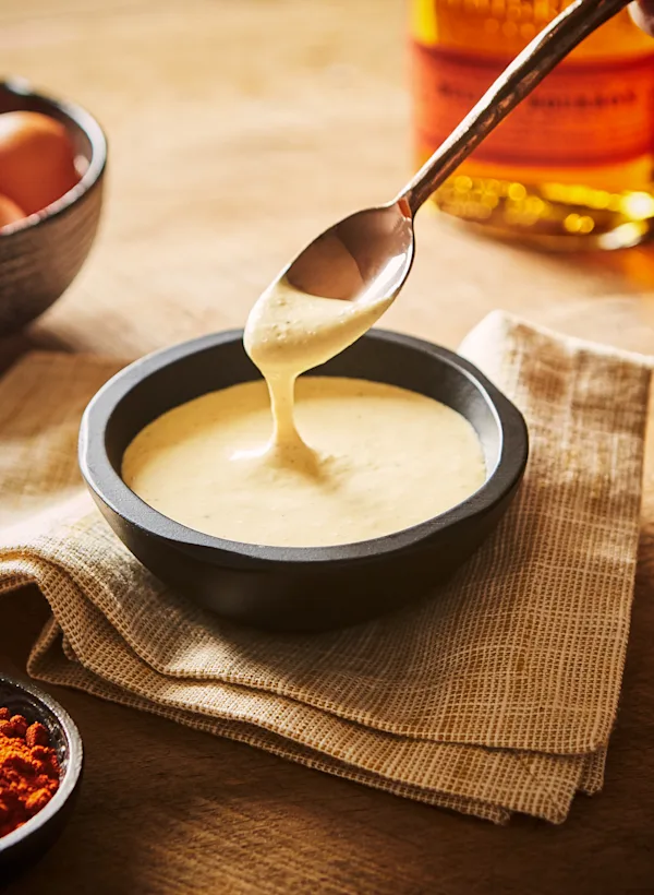 A black dish containing a cream colour sauce sits on a table. A spoon dips into it. Also on the table is a bottle of bourbon, a bowl of eggs and a bowl of spices.