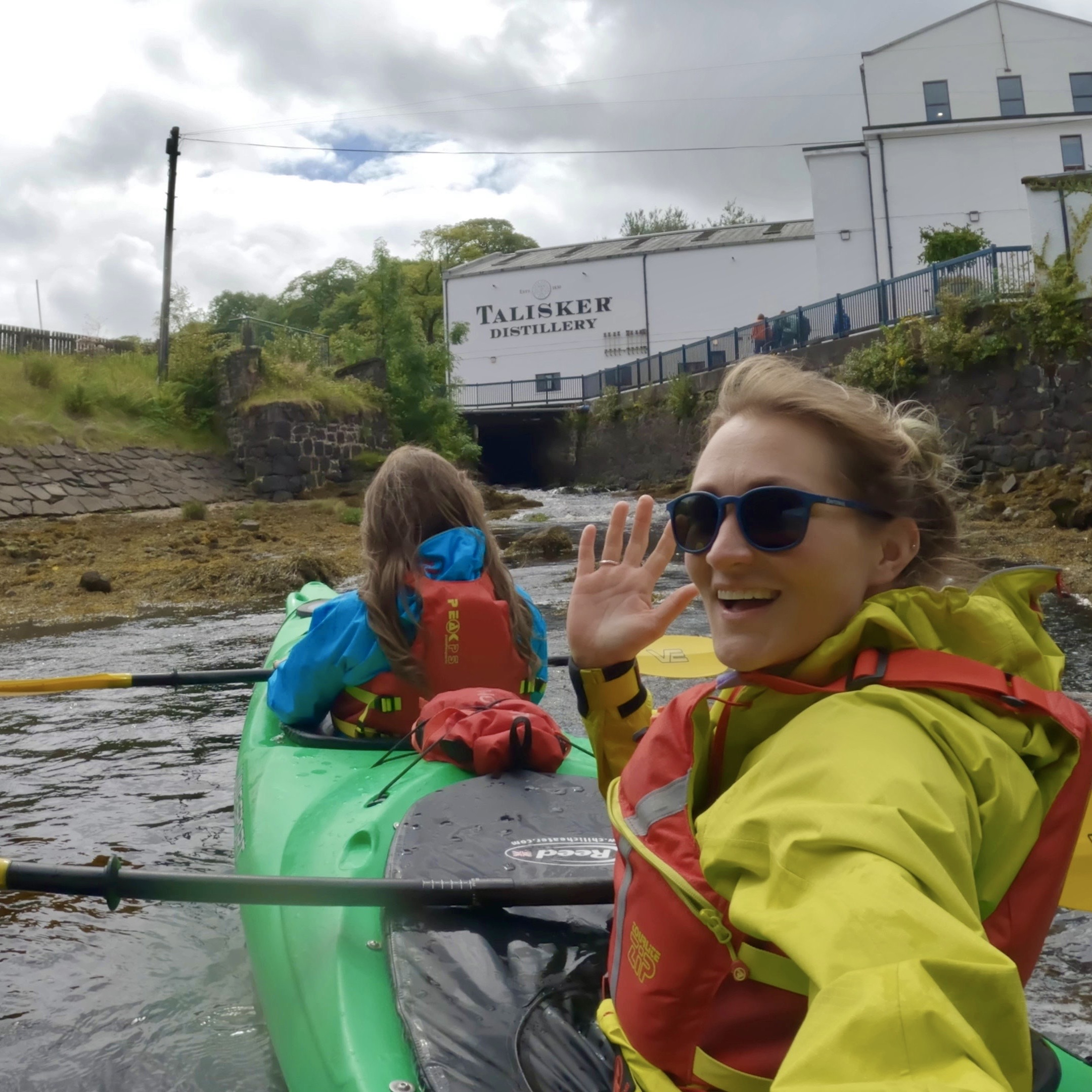 Two women sit in a kayak on the water in front of Talisker distillery. One faces the camera, waving.