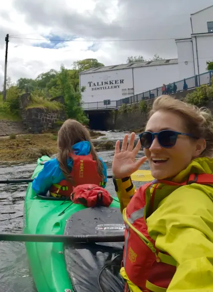 Twee vrouwen zitten in een kajak op het water voor de Talisker distilleerderij. De ene kijkt zwaaiend naar de camera.