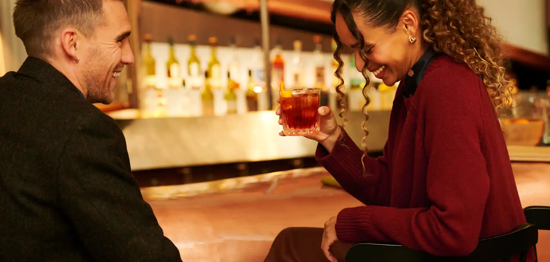 Ein Mann und eine Frau sitzen an einer Bar, sie hält einen Whisky-Cocktail 