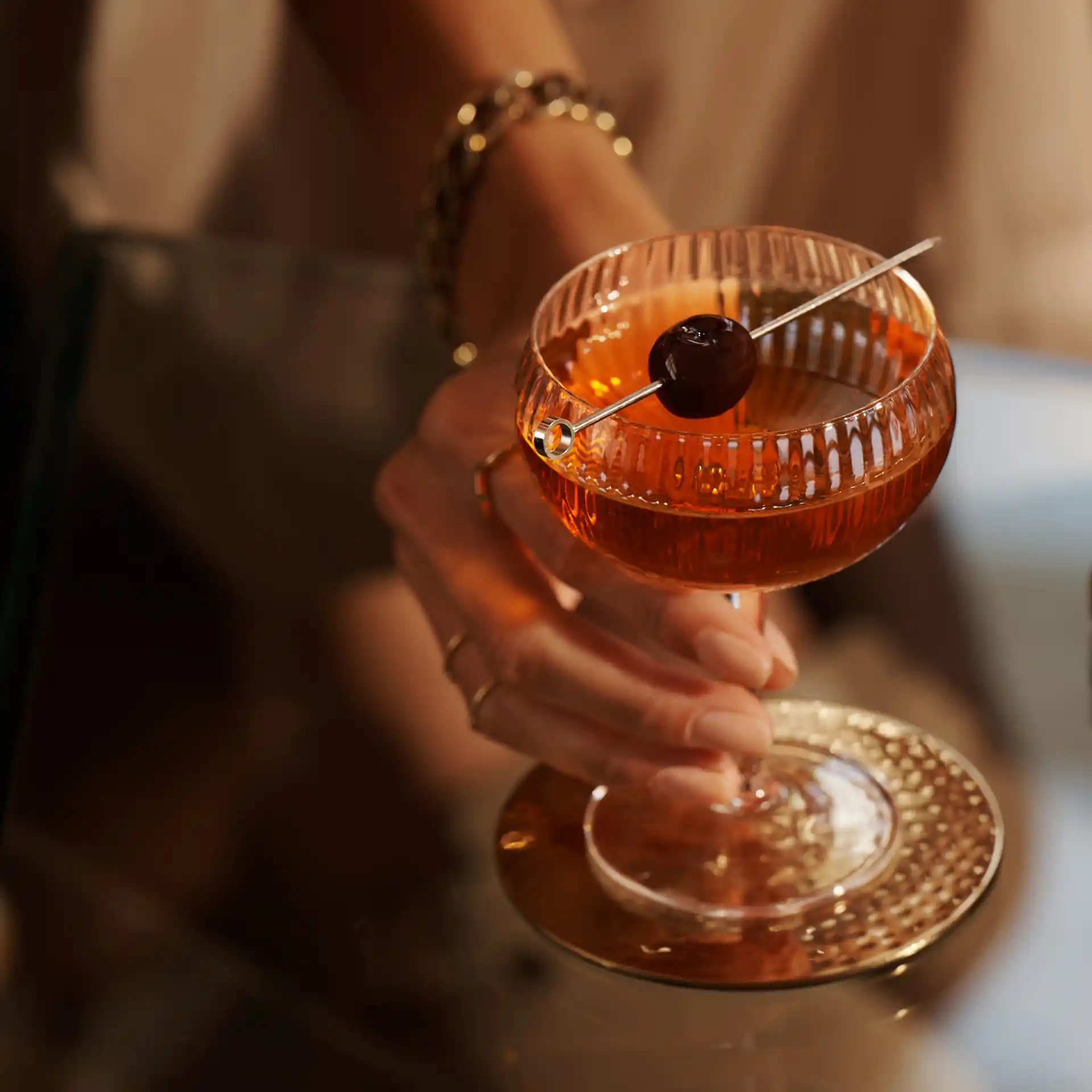 A Manhattan cocktail sits on a glass table, with a skewed maraschino cherry sitting over the rim of the coupe glass. A person holds the coupe by the stem.