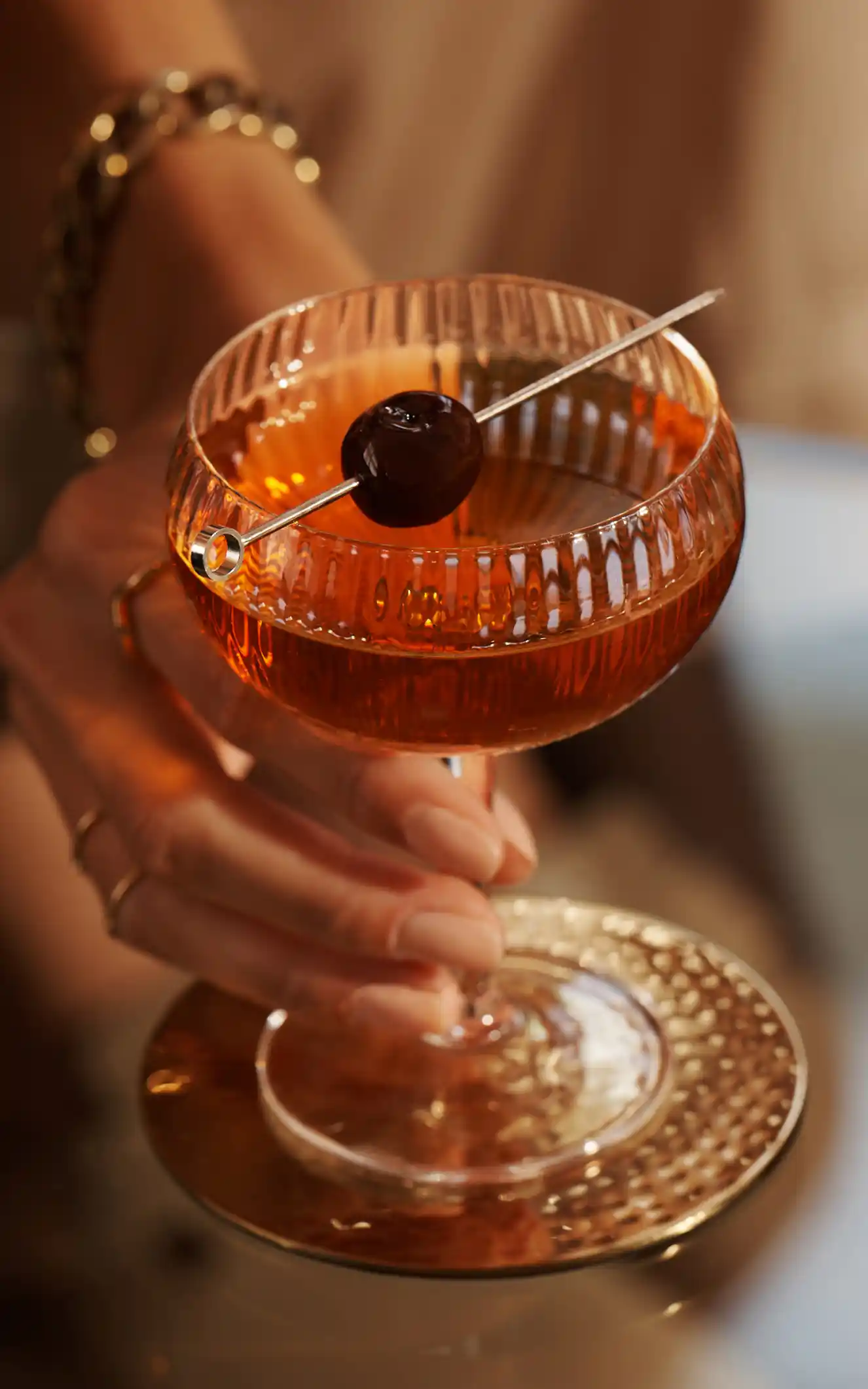 Een Manhattan-cocktail staat op een glazen tafel, met een scheve maraschino-kers over de rand van het coupeglas. Iemand houdt de coupe bij de steel vast.