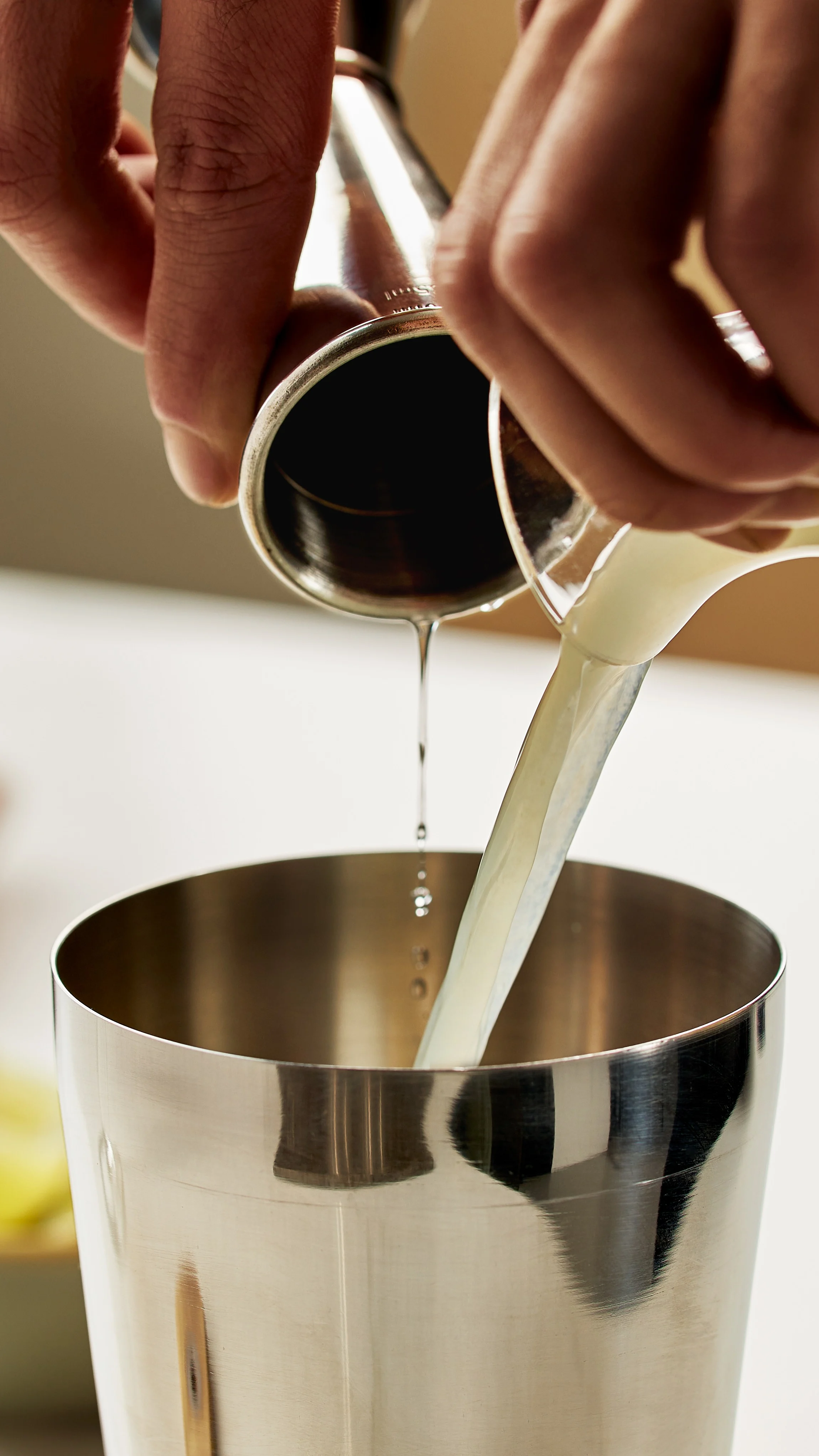 A jigger and a small jug pour alcohol and lime into a metal cocktail shaker.  