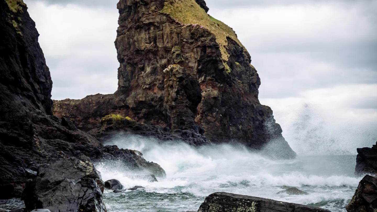 : A large, rocky cliff is surrounded by smaller cliffs and rocks. A wild sea crashes against the bottom of the cliff and sends spray up into the air.