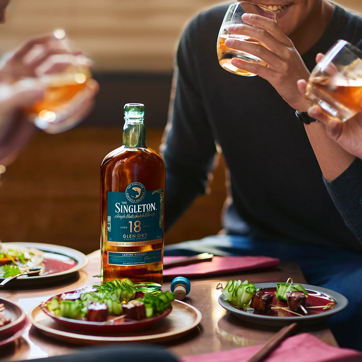 The Singleton of Glen Ord 18 Year Old Single Malt Scotch Whisky Bottle on a table with some snacks in the middle of a group of people enjoying