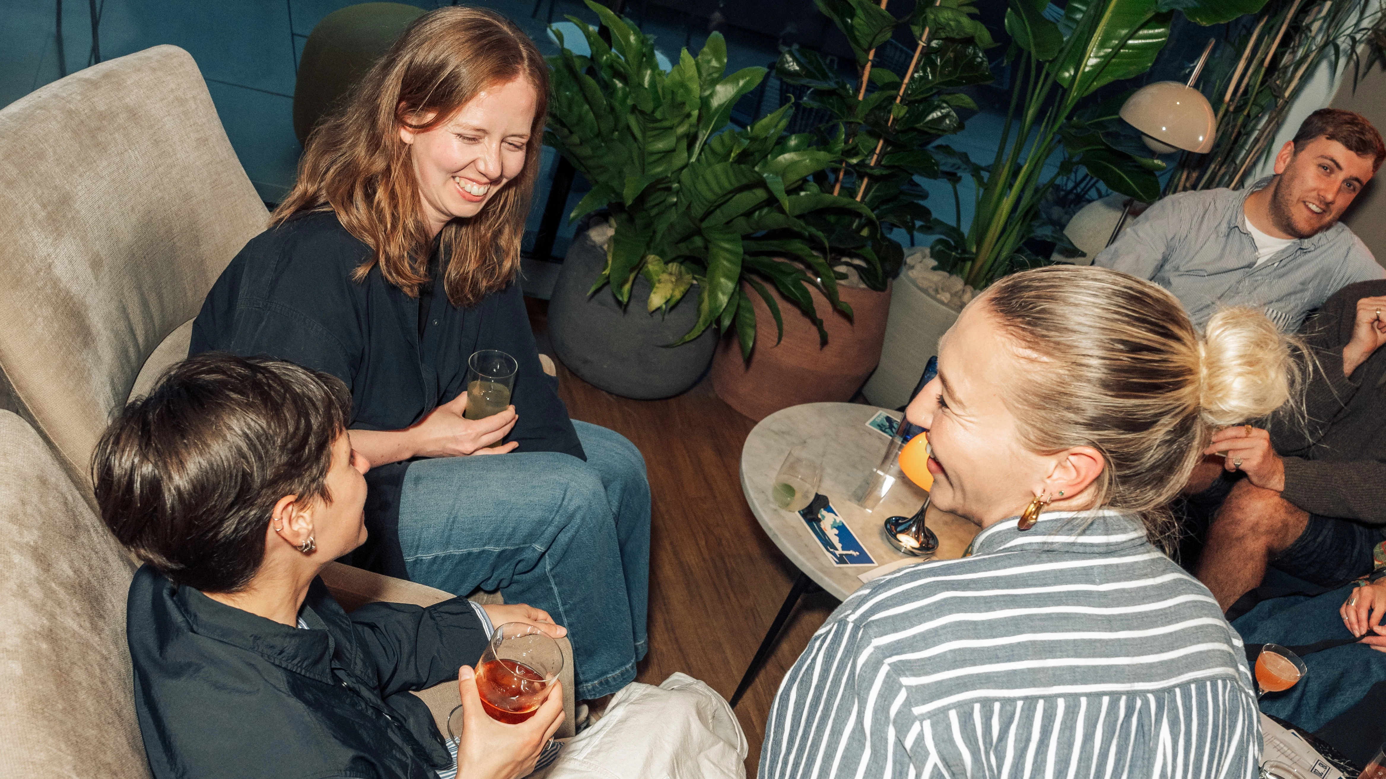 A group of three female friends drinking cocktails amd sat in chairs laughing together
