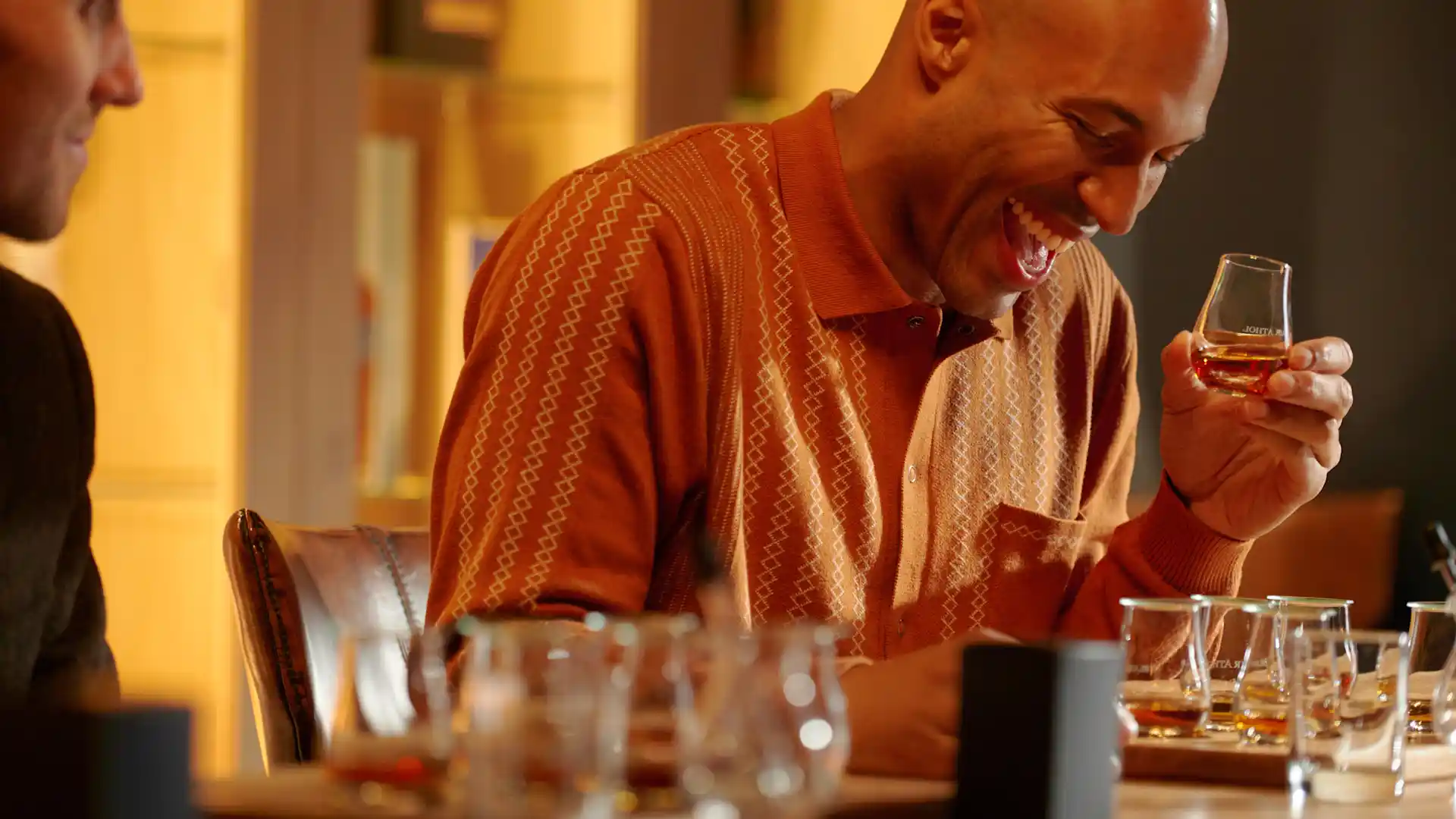 Two male friends sit at a table enjoying a whisky tasting experience at Blair Athol distillery. One is laughing while holding a tasting glass