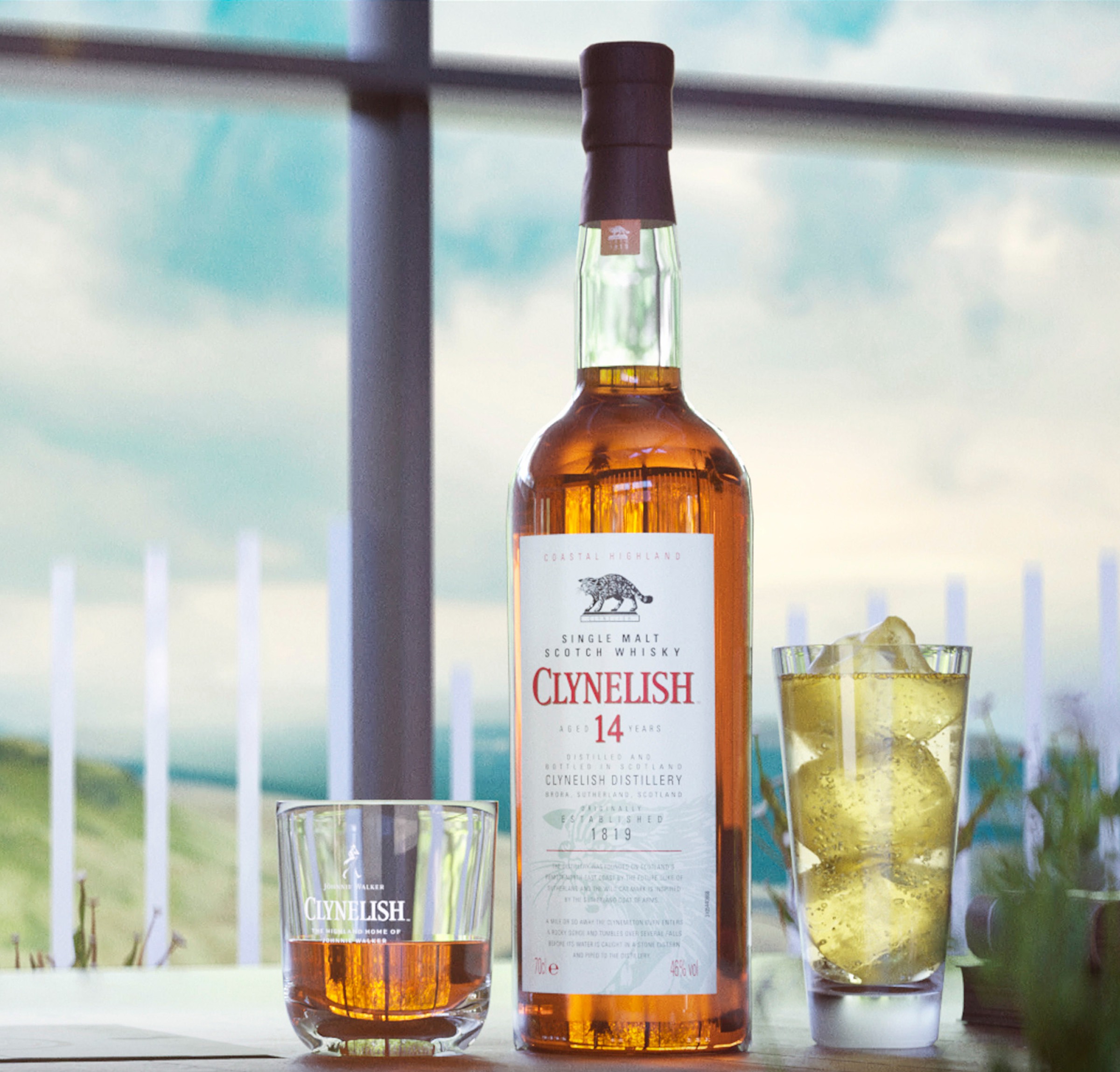 A bottle of Clynelish 14 year old whisky sits on a table next to a rocks glass containing whisky and a highball glass containing a light yellow cocktail and ice cubes.