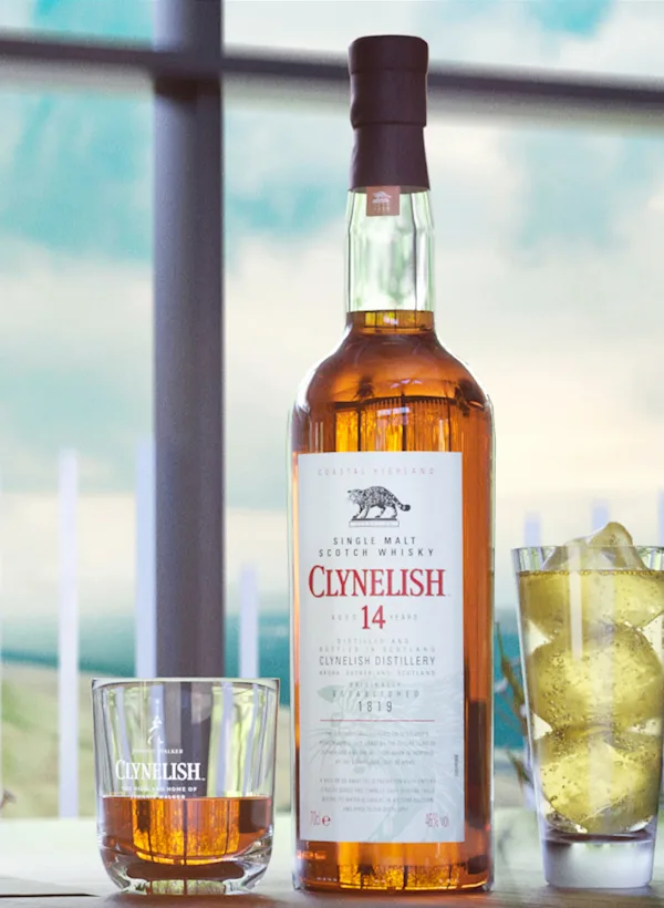 Eine Flasche Clynelish 14 Jahre alter Whisky steht auf einem Tisch neben einem Rocks-Glas mit Whisky und einem Highball-Glas mit einem hellgelben Cocktail und Eiswürfeln.