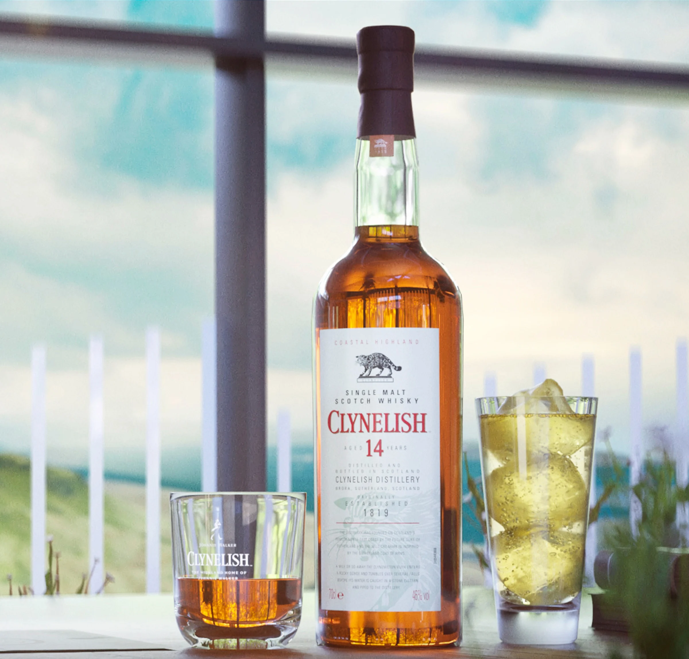 A bottle of Clynelish 14 year old whisky sits on a table next to a rocks glass containing whisky and a highball glass containing a light yellow cocktail and ice cubes.