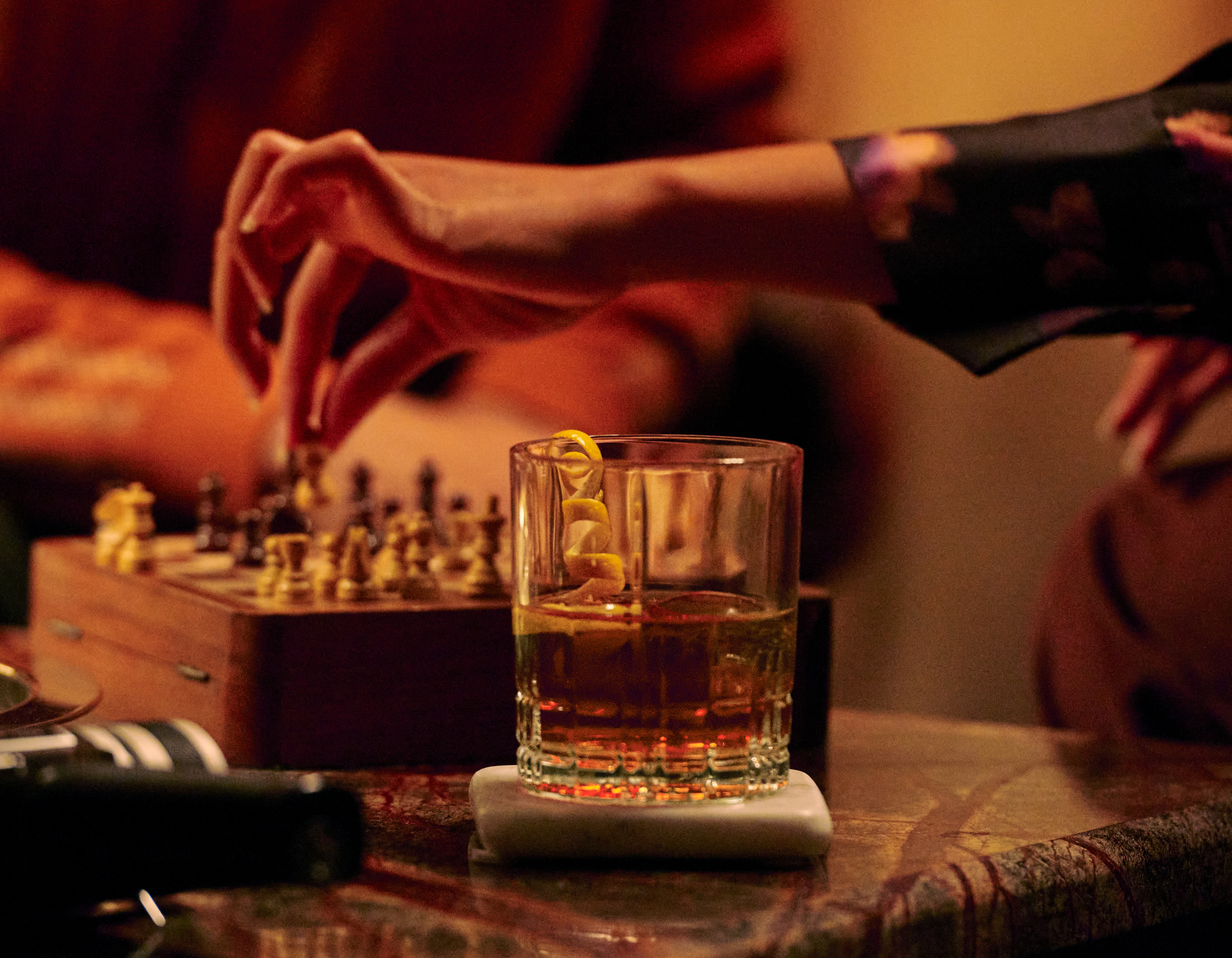 A rocks glass with a pouring of whisky, as people play chess in the background
