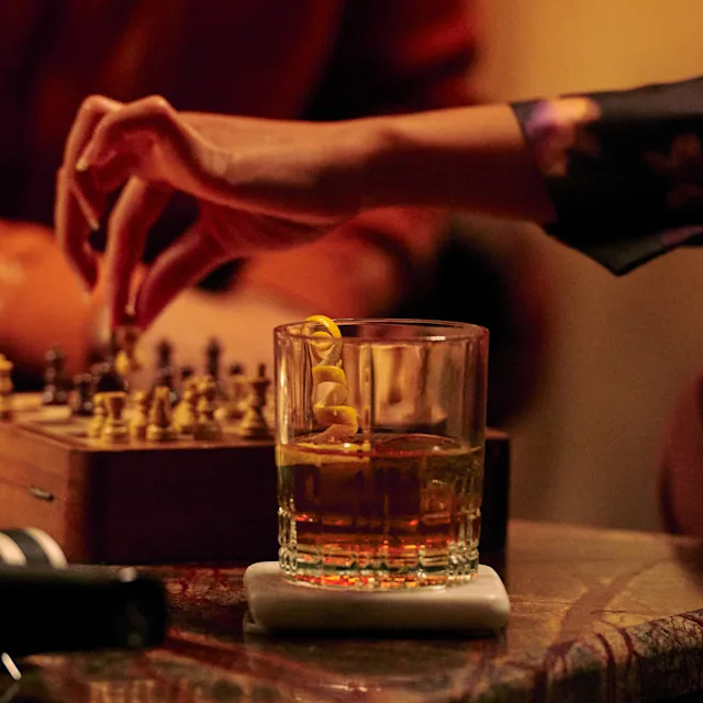 A rocks glass with a pouring of whisky, as people play chess in the background