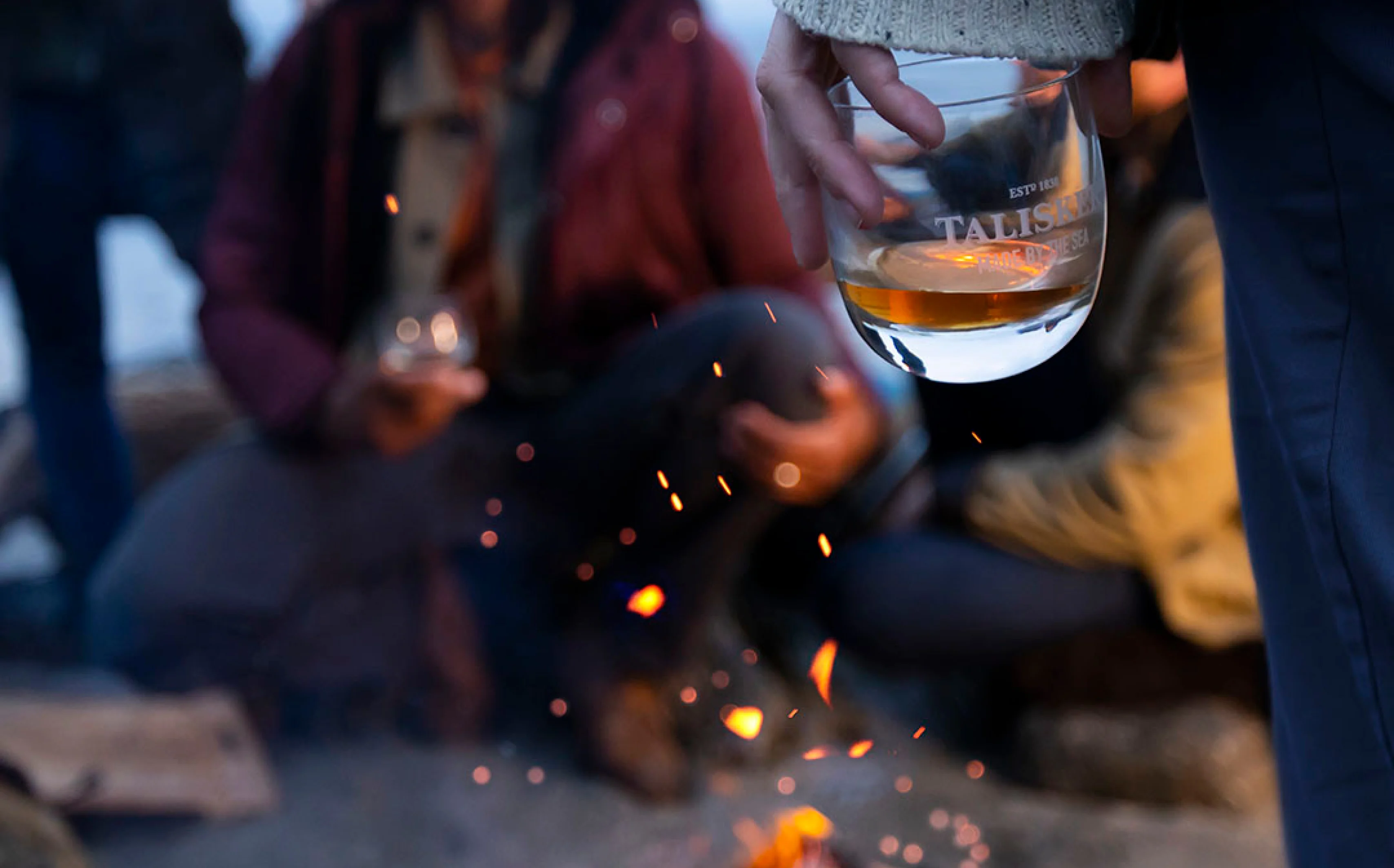 A group of people sit around a small bonfire, holding Talisker-branded rocks glasses containing whisky.