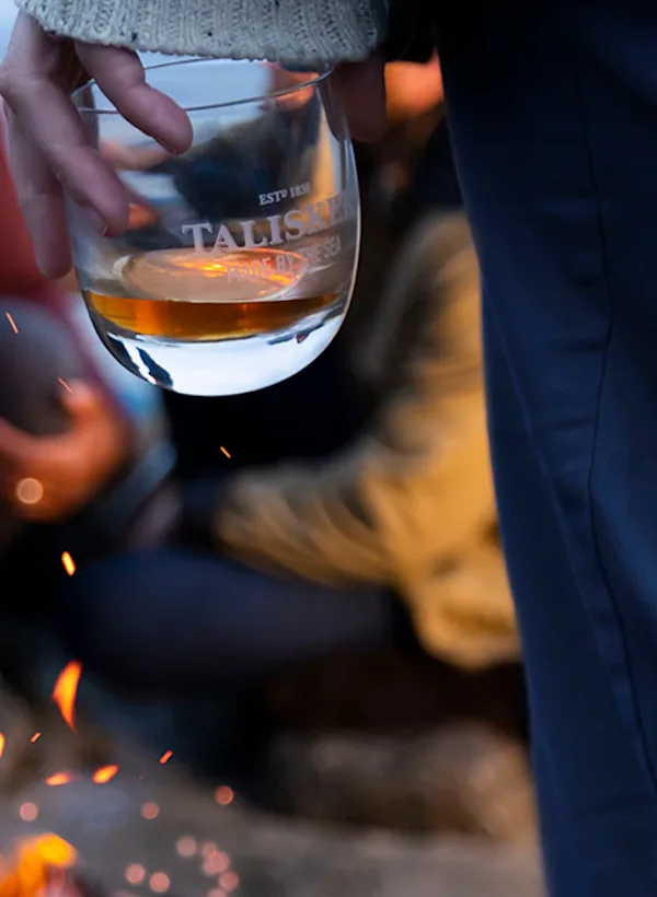 Ontdek Een groep mensen zit rond een klein kampvuur en houdt glazen met whisky van het merk Talisker vast.