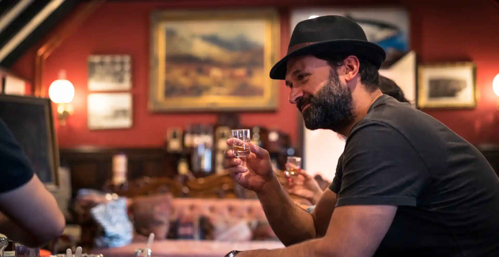 Een man zit met een groep mensen en houdt een klein glas whisky vast. Hij glimlacht naar iemand buiten de camera. Voor hem staat een lage tafel met verschillende whiskyflessen, glazen en pipetten.