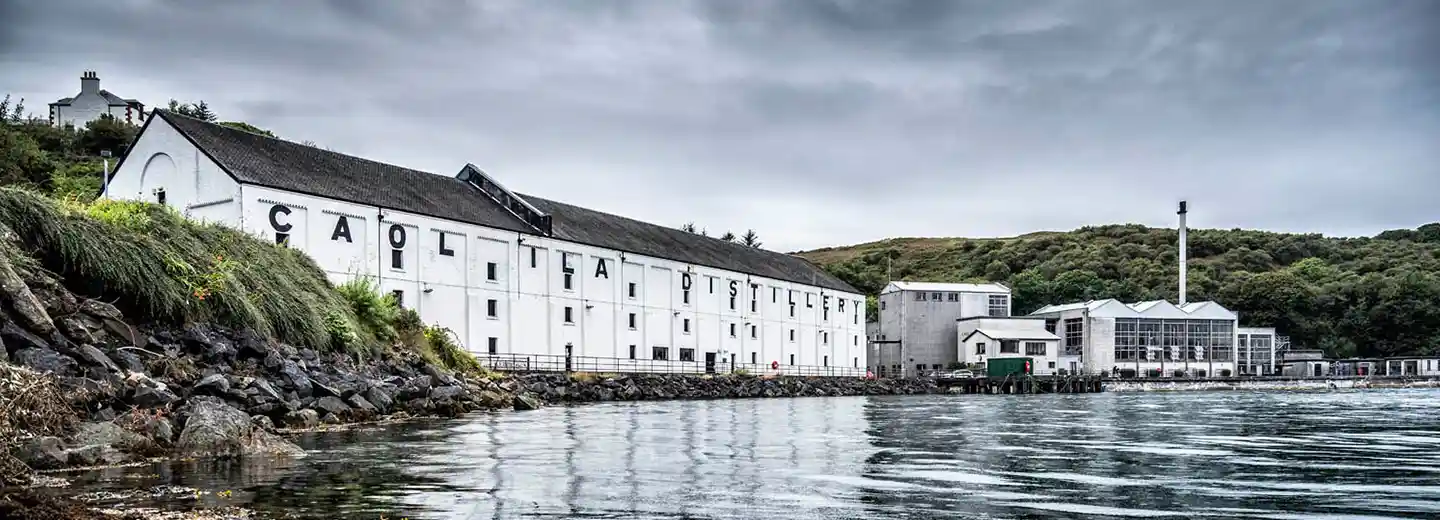 The white building of Caol Ila distillery is shown sitting on a body of water, surrounded by a rocky shore and mountains in the background. 