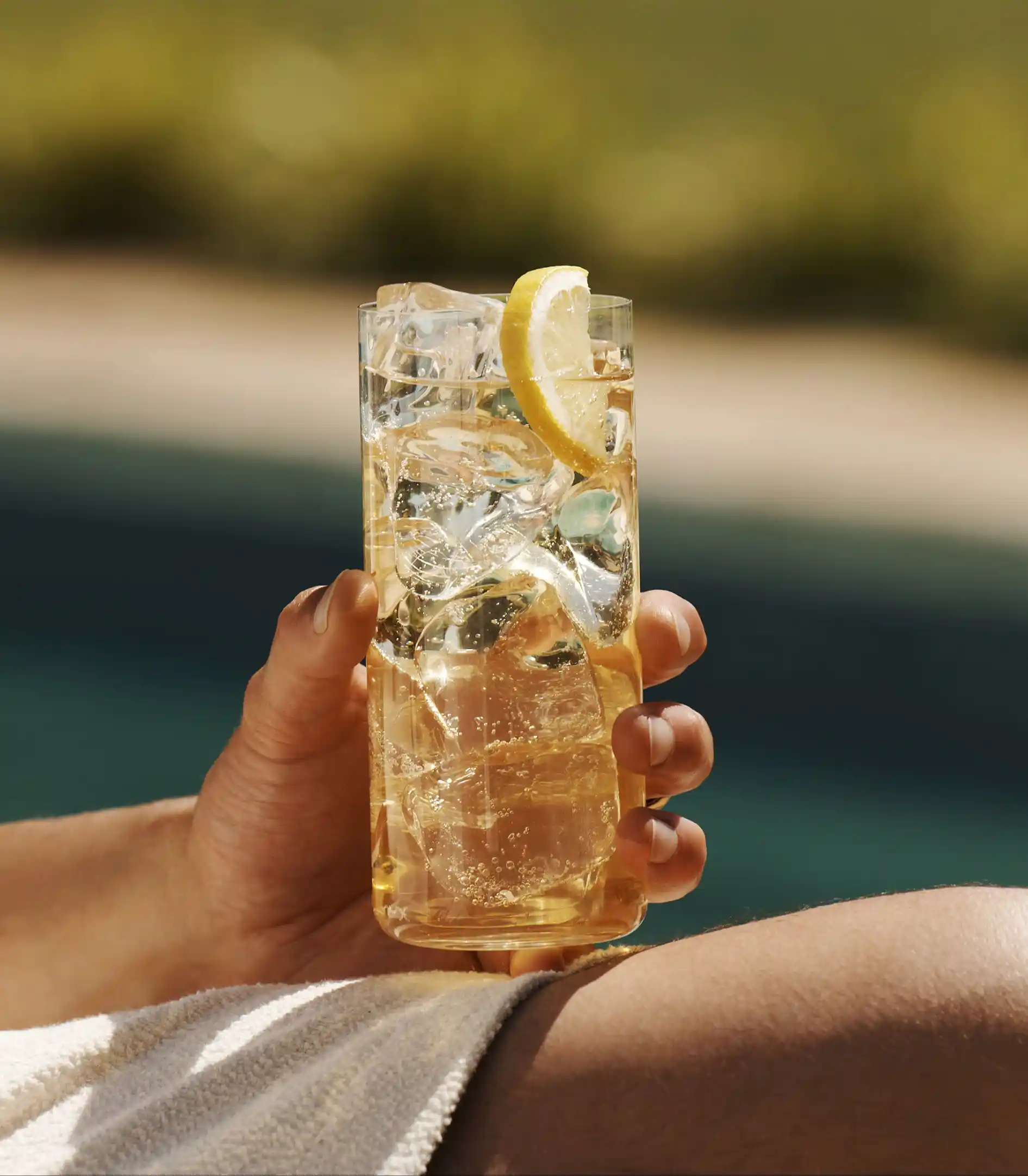 A man holds a whisky highball on his knee while sat next to a pool