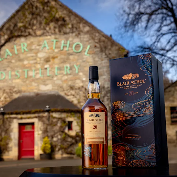 A bottle of Blair Athol 20-year-old Highland Single Malt Scotch Whisky with its packaging box displayed in front of the historic Blair Athol Distillery building.
