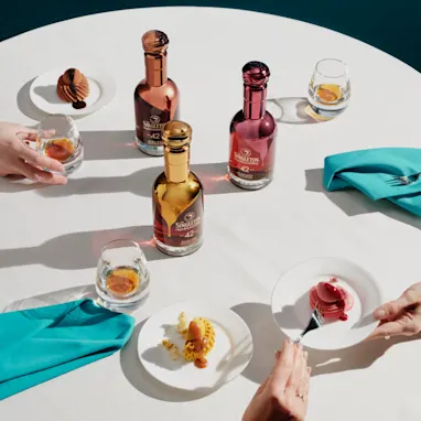 Three bottles of The Singleton Gourmand Collection sit on a white tablecloth, surrounded by three plates of cakes with rocks glasses containing whisky.